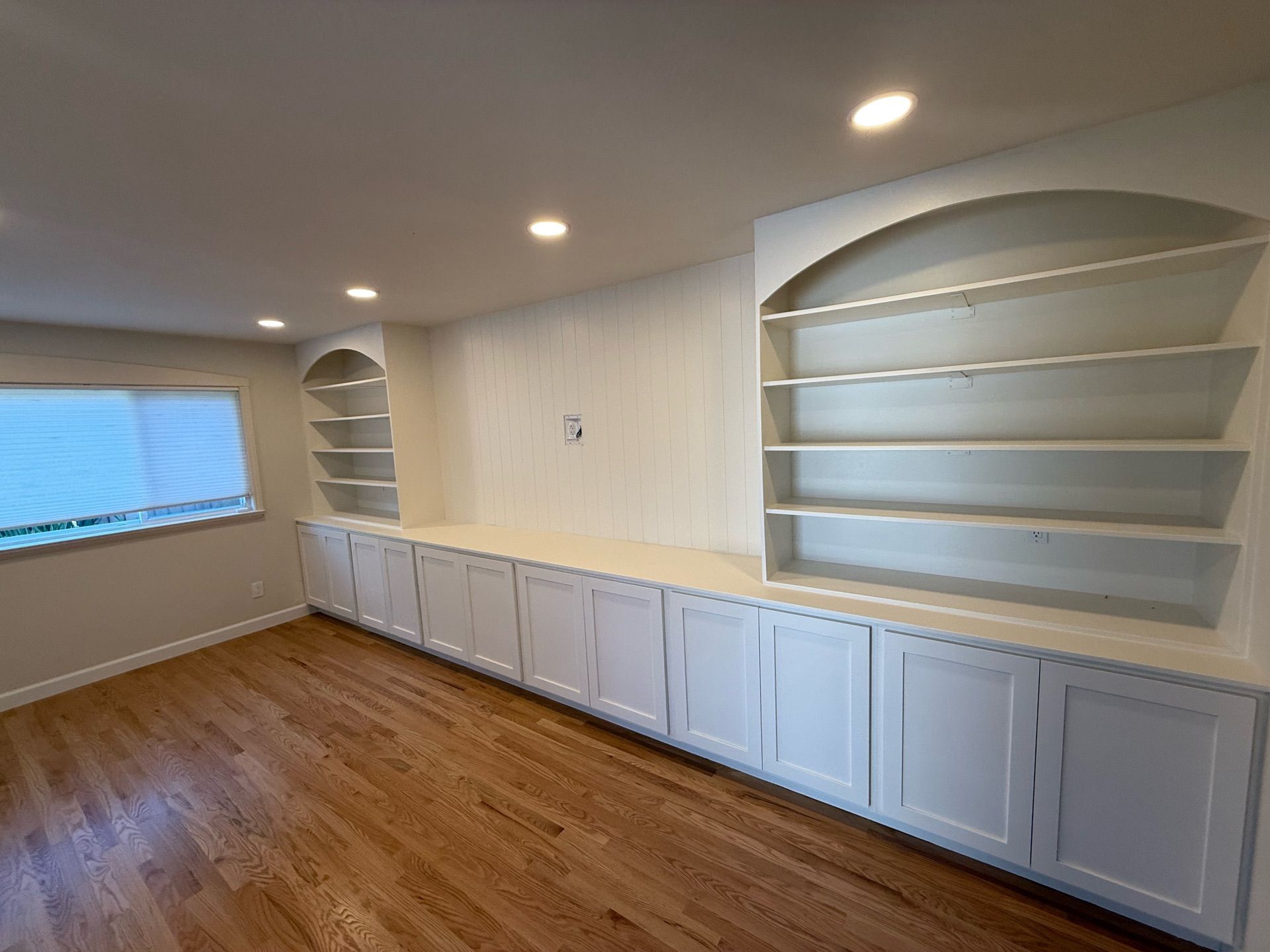 Built-in white cabinets and bookshelves with recessed lighting in a room with hardwood floors and a window.