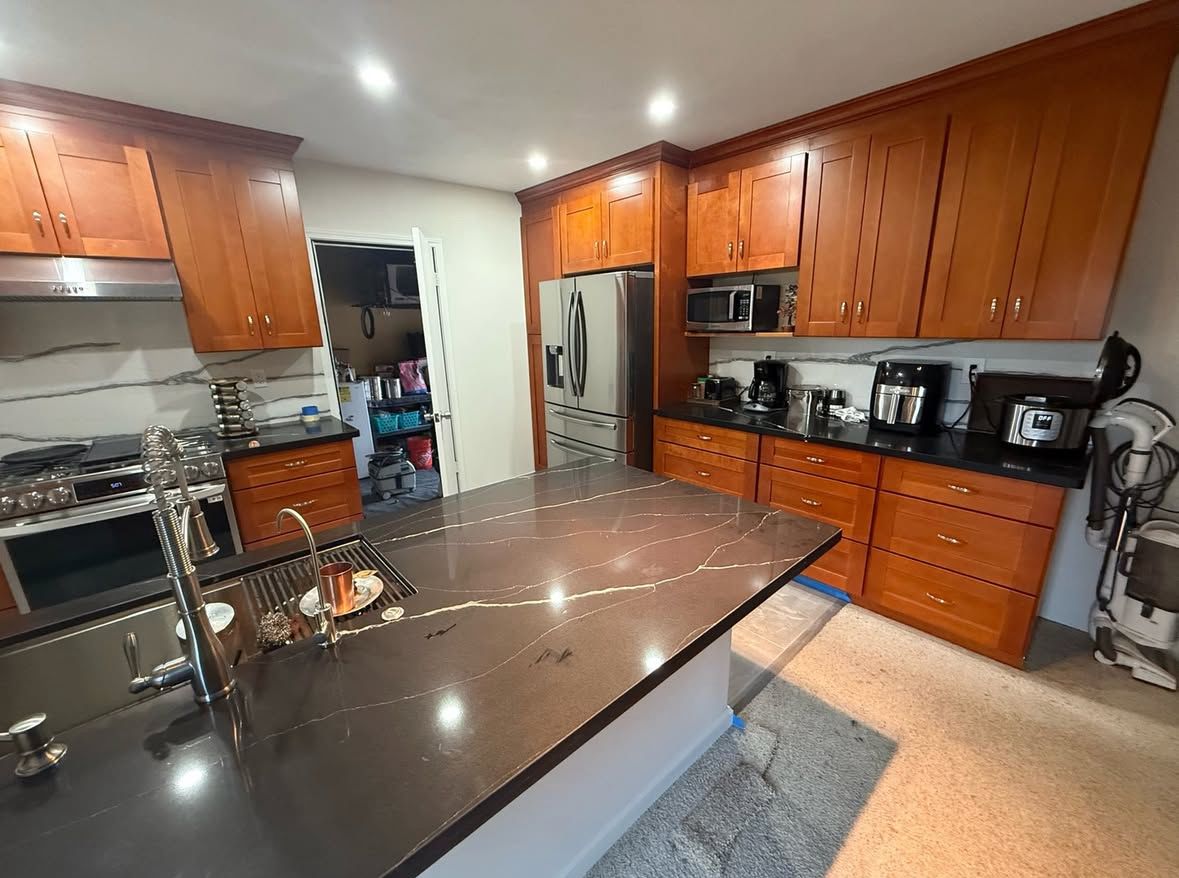 Kitchen with brown cabinets, dark countertop island, and stainless steel appliances. A doorway leads to a cluttered pantry.