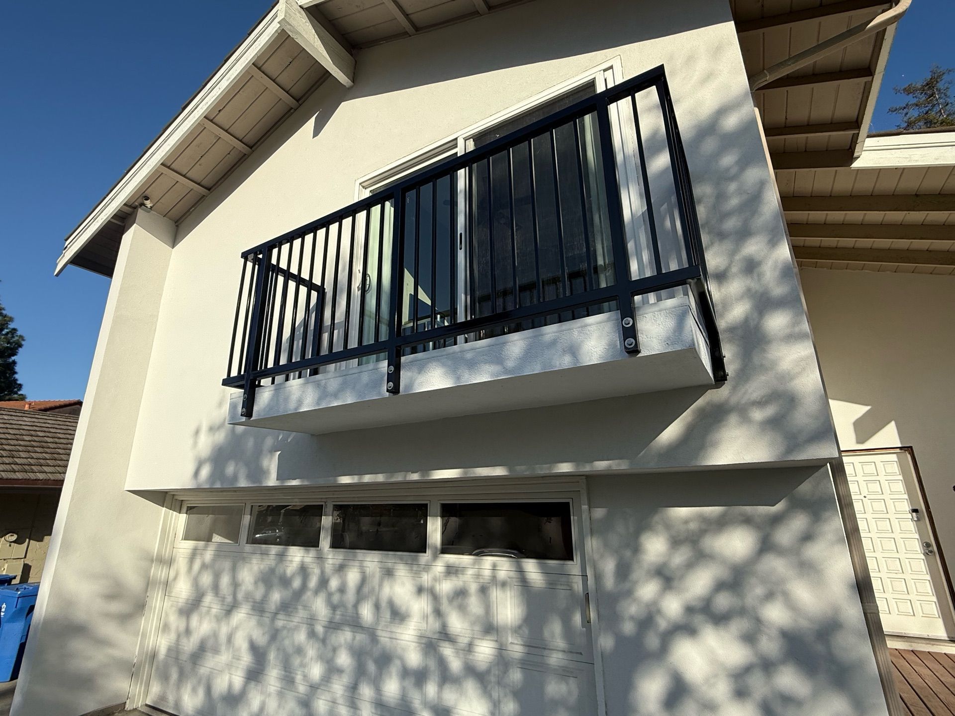 Exterior of a light-colored house with a small balcony featuring black railing above a garage. Clear blue sky.