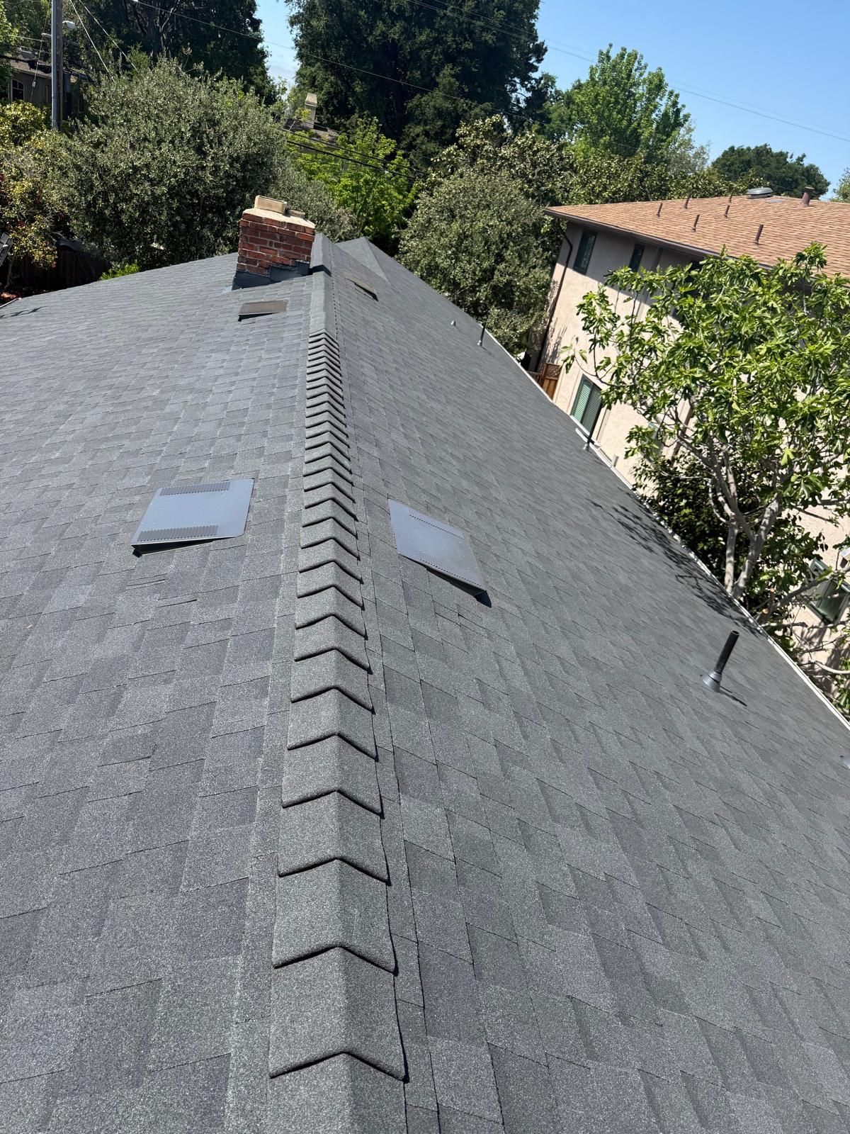 Overhead view of a dark shingled roof, showing the ridge, two vent covers, and a brick chimney.