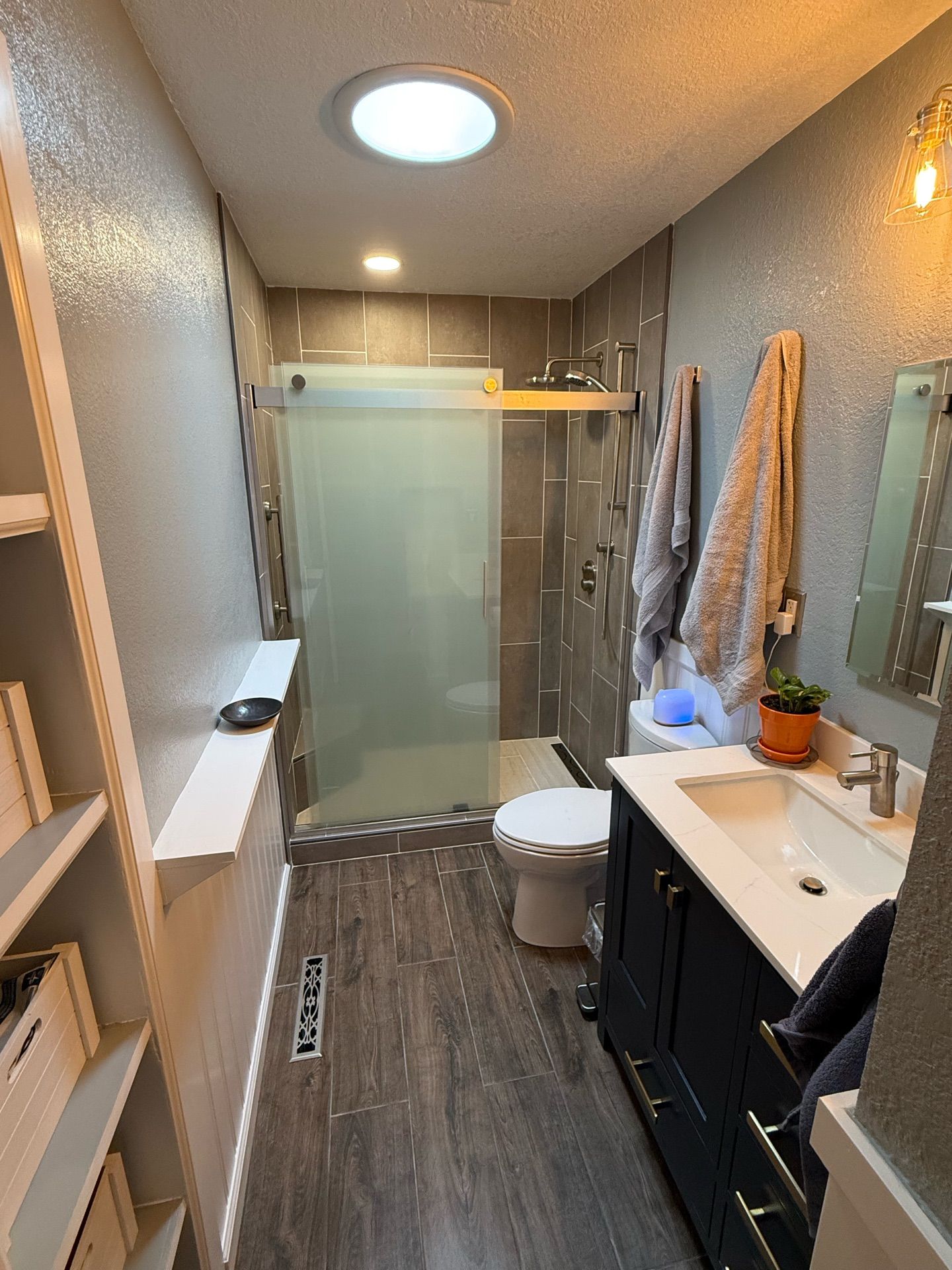 A modern bathroom with a shower, vanity, and toilet. The space has gray tile, wood-look flooring, and a skylight.