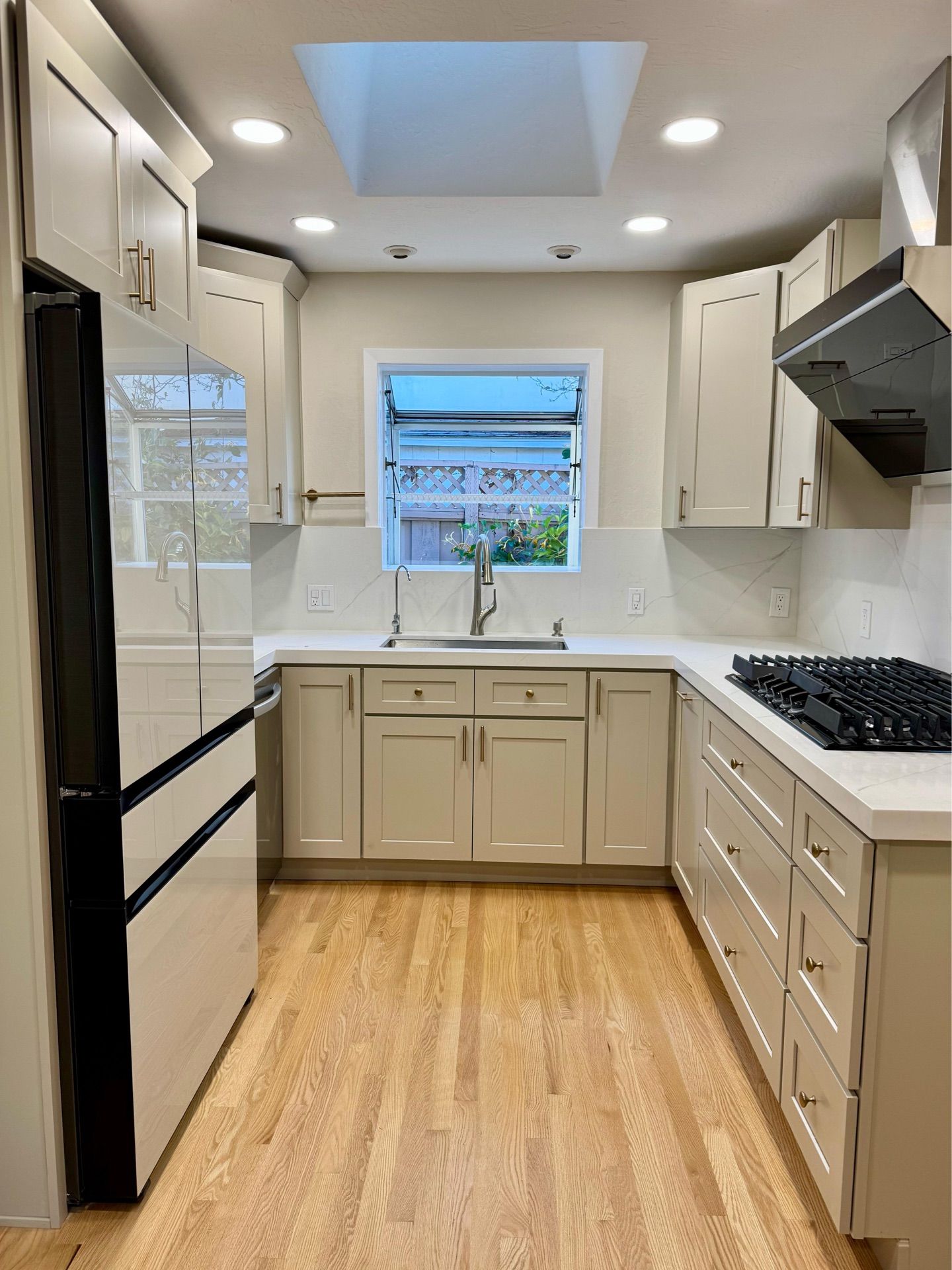A renovated kitchen with light-colored cabinets, stainless steel appliances, and hardwood floors. A window and skylight provide natural light.