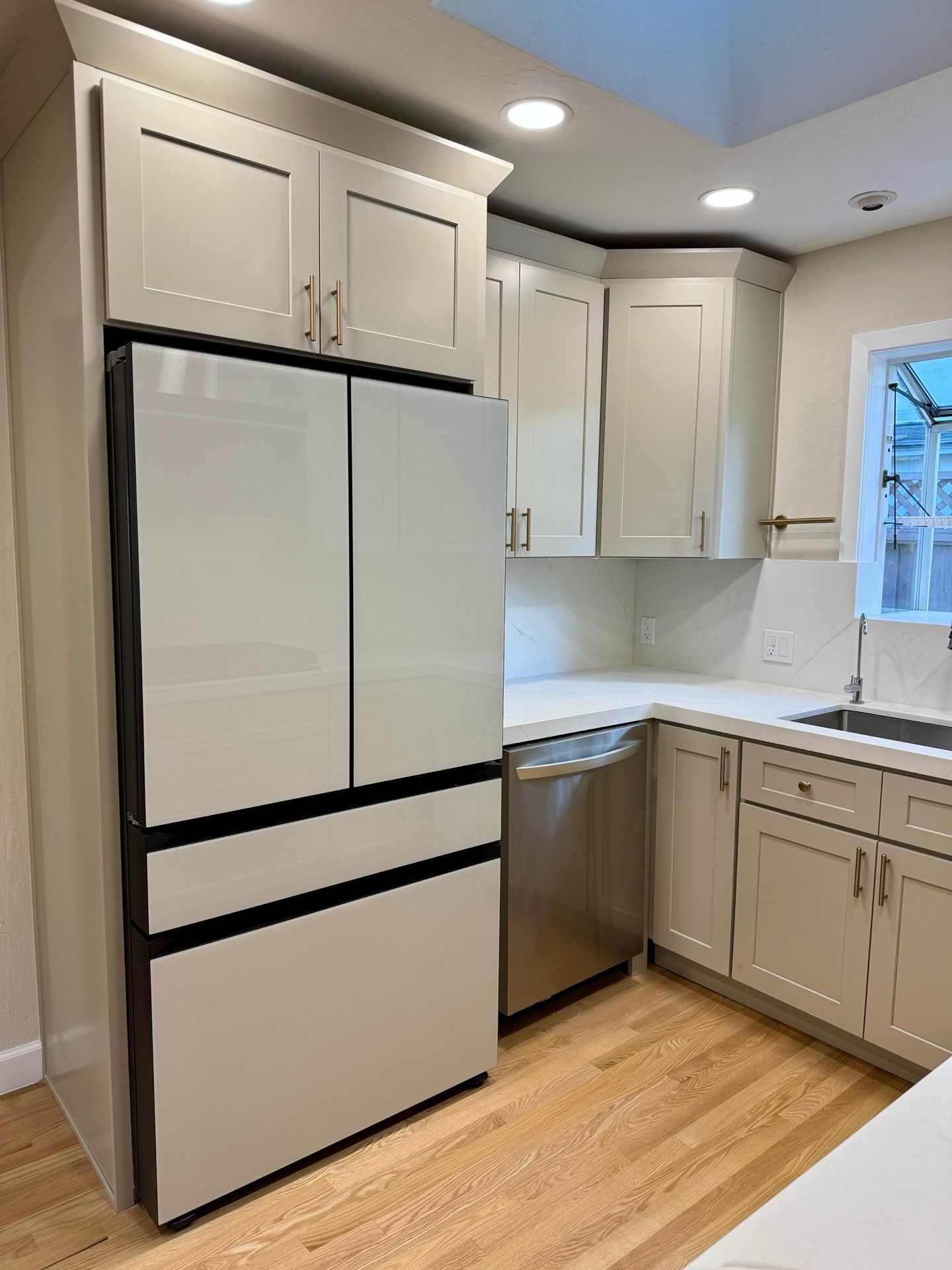 A modern kitchen with light gray cabinets, white countertops, and a white refrigerator. The room has a window and light hardwood floors.