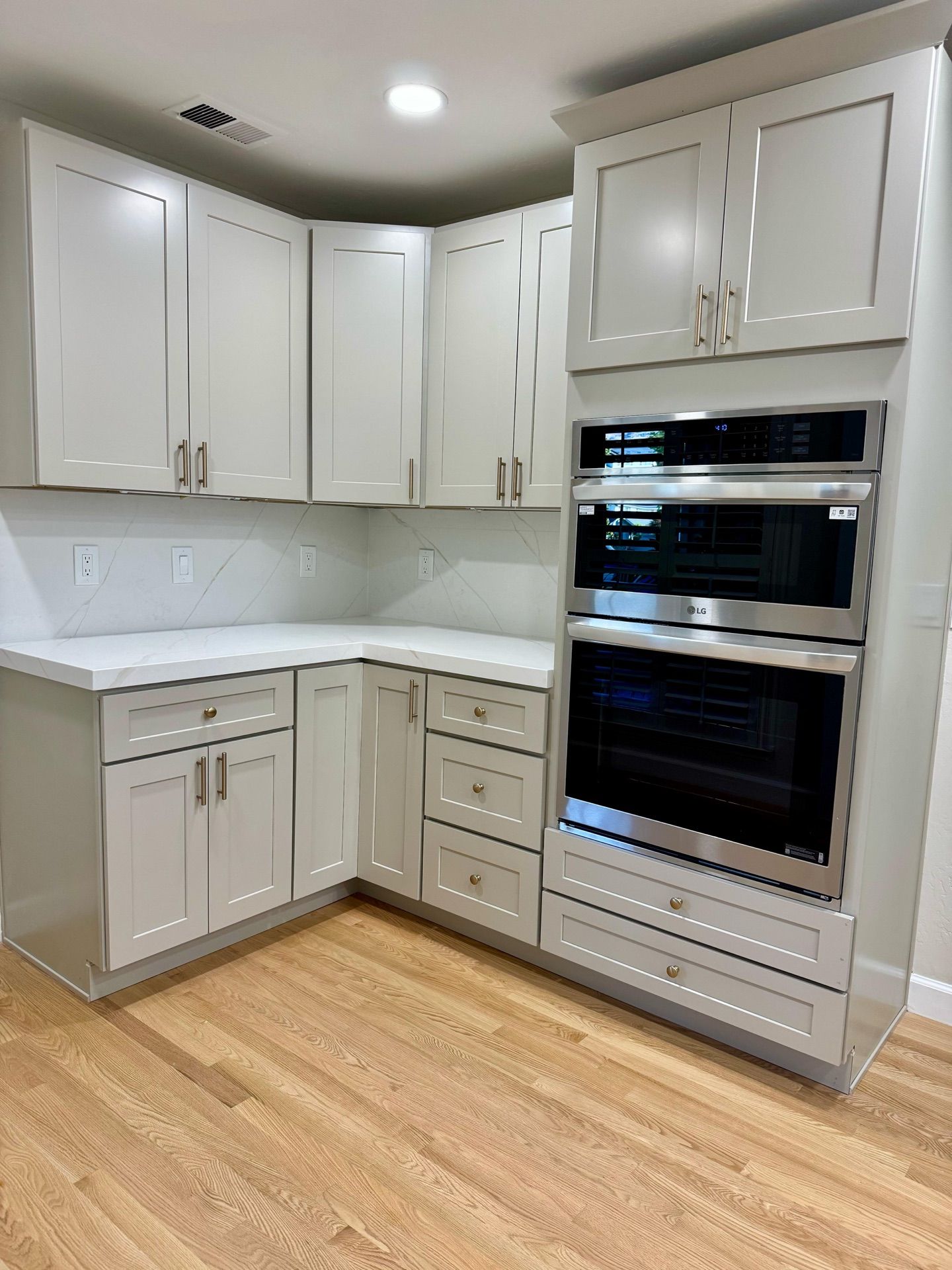 Light gray kitchen cabinets with white countertops and a built-in stainless steel oven, on a hardwood floor.