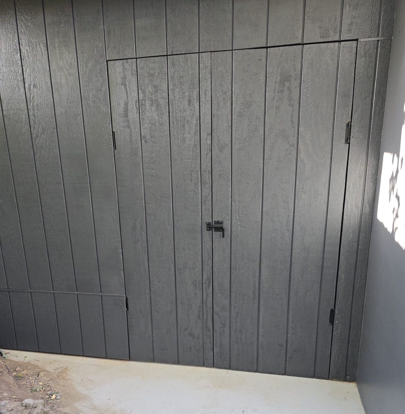 Dark gray wooden door with a small access door, set against a gray wall in a sunny outdoor space.