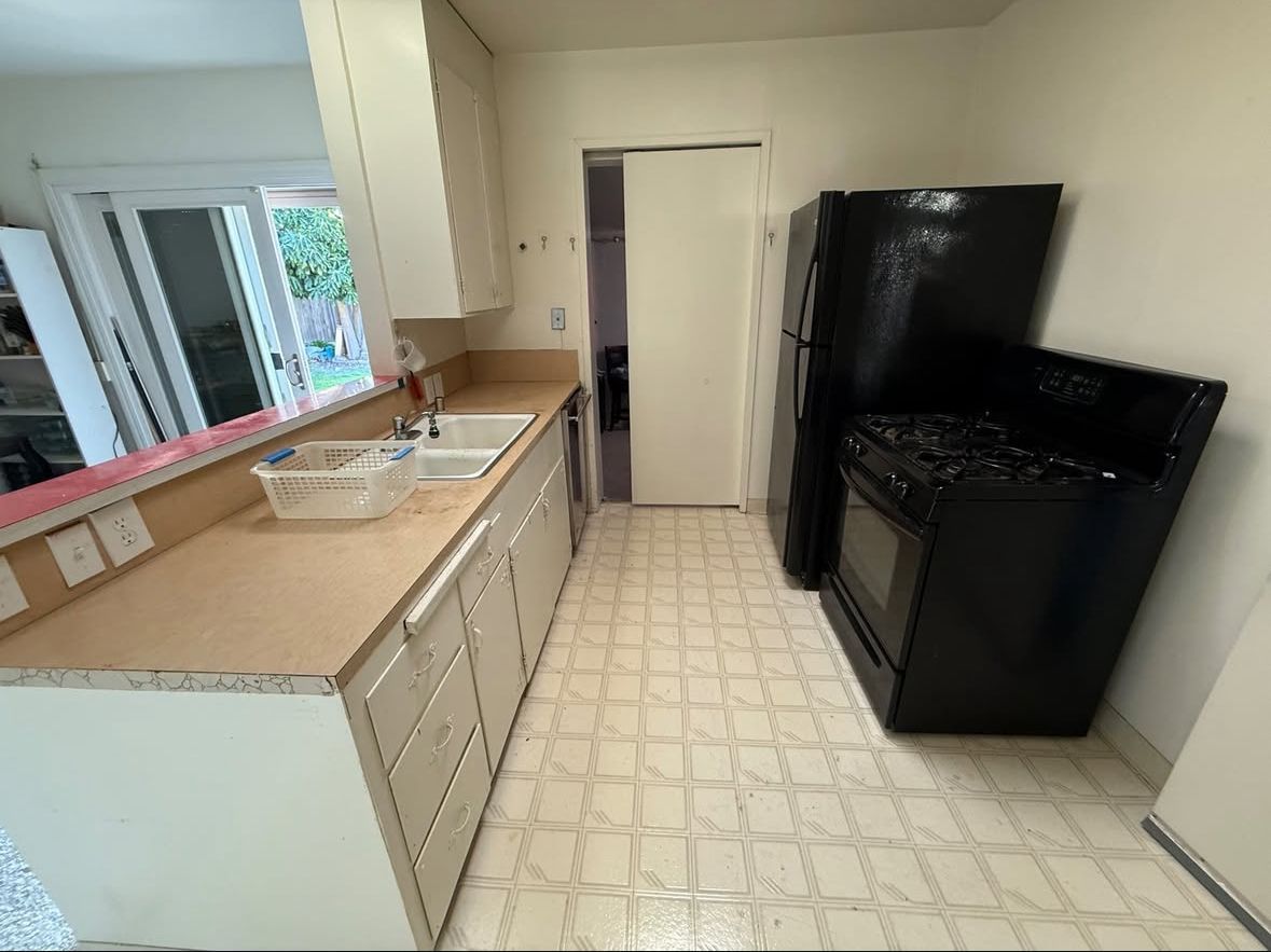 A small, cluttered kitchen with white cabinets, a beige countertop, and a black refrigerator and stove. A doorway leads to another room.