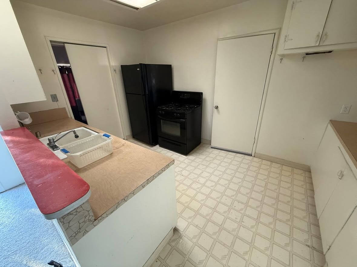 A small kitchen with white cabinets, a black refrigerator and stove, and beige countertops. A sink is in the foreground.
