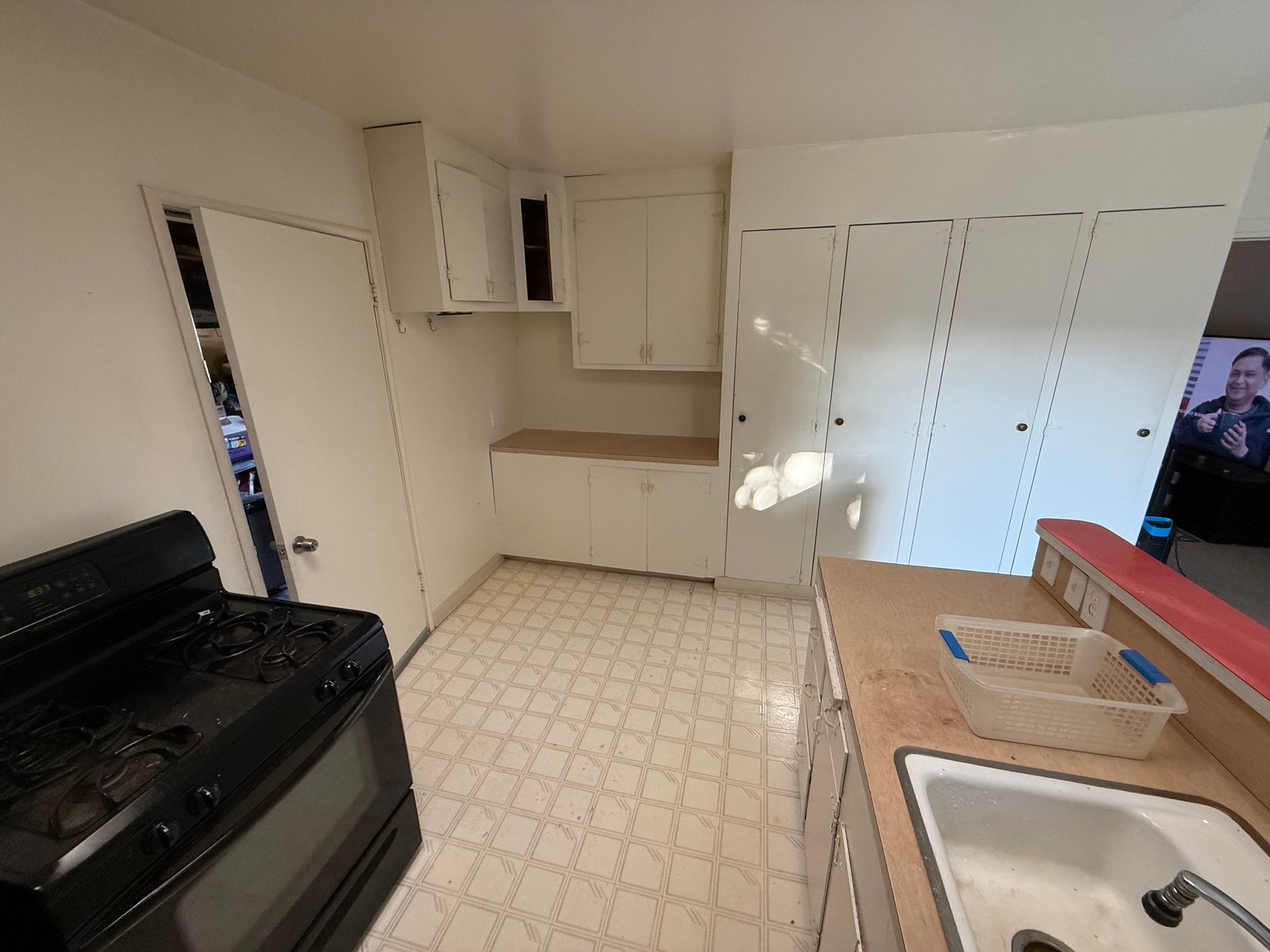 A small kitchen with white cabinets, appliances, and a patterned floor. An open door reveals a cluttered space.