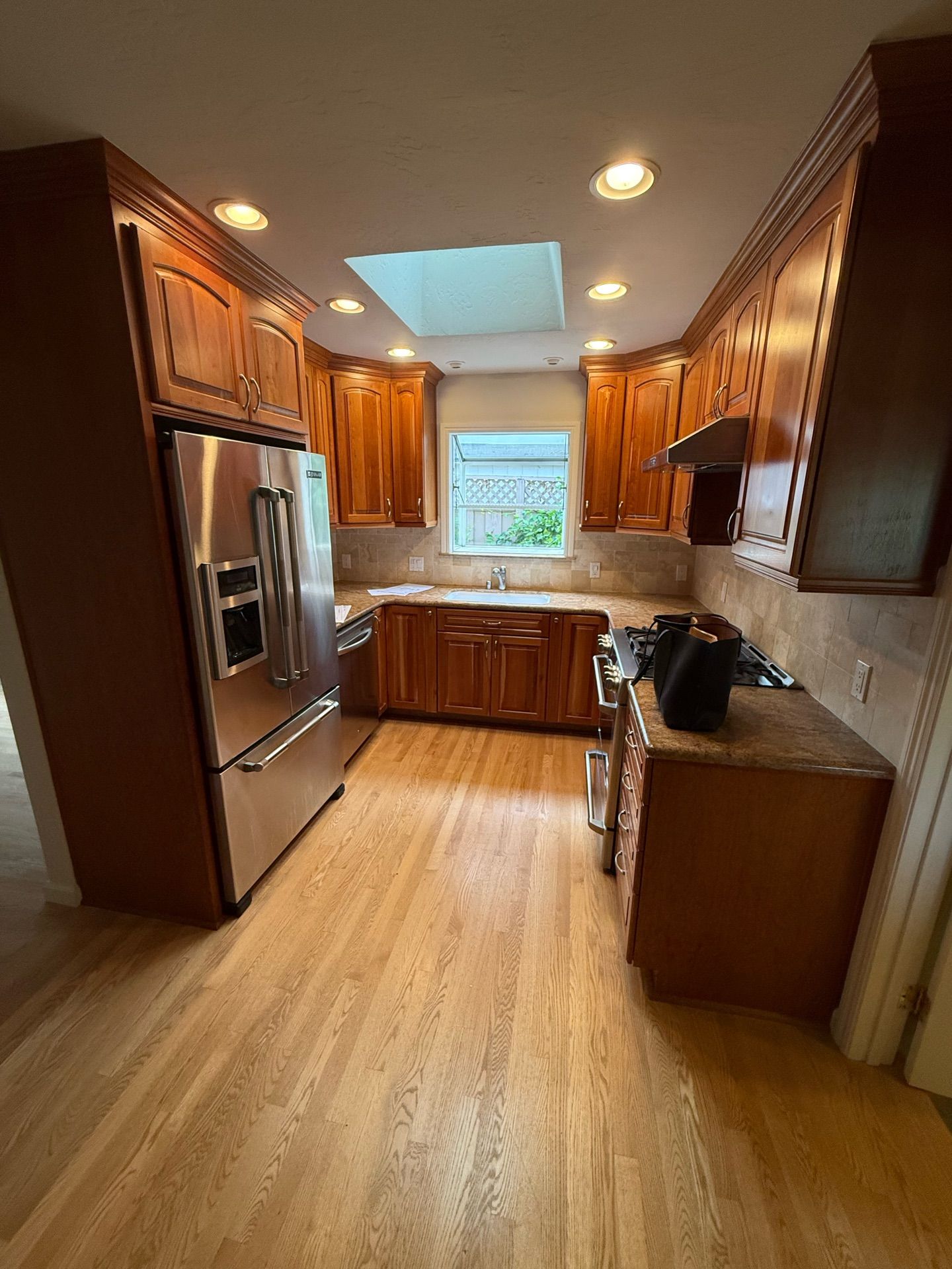 A kitchen with wood cabinets and flooring, stainless steel refrigerator, and skylight, with a window over a sink.