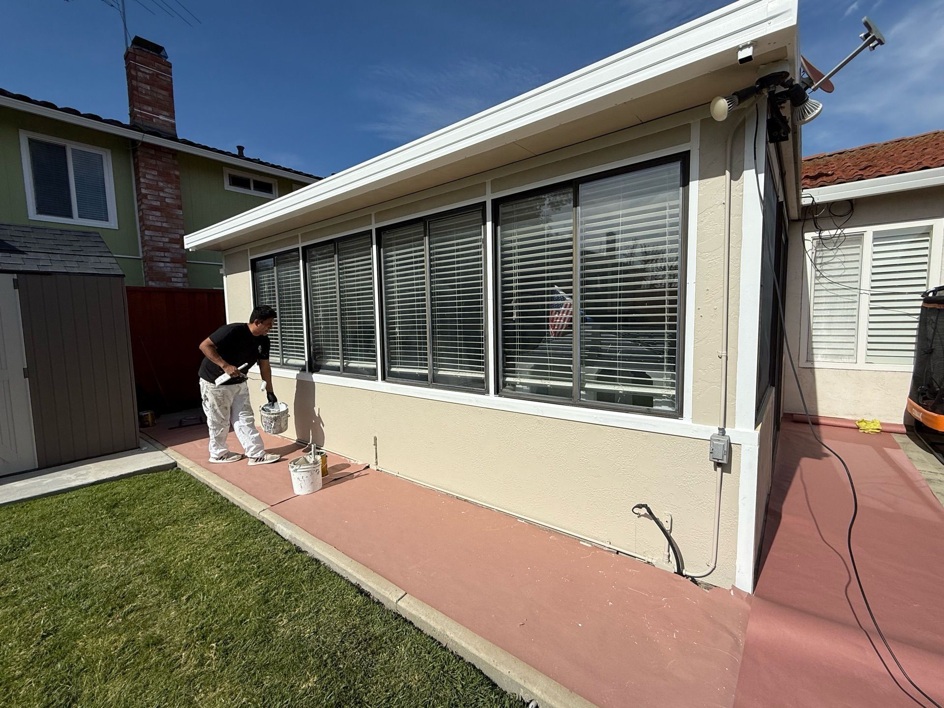 A person paints the exterior wall of a house. The house has multiple windows with blinds. The setting is outdoors on a sunny day.