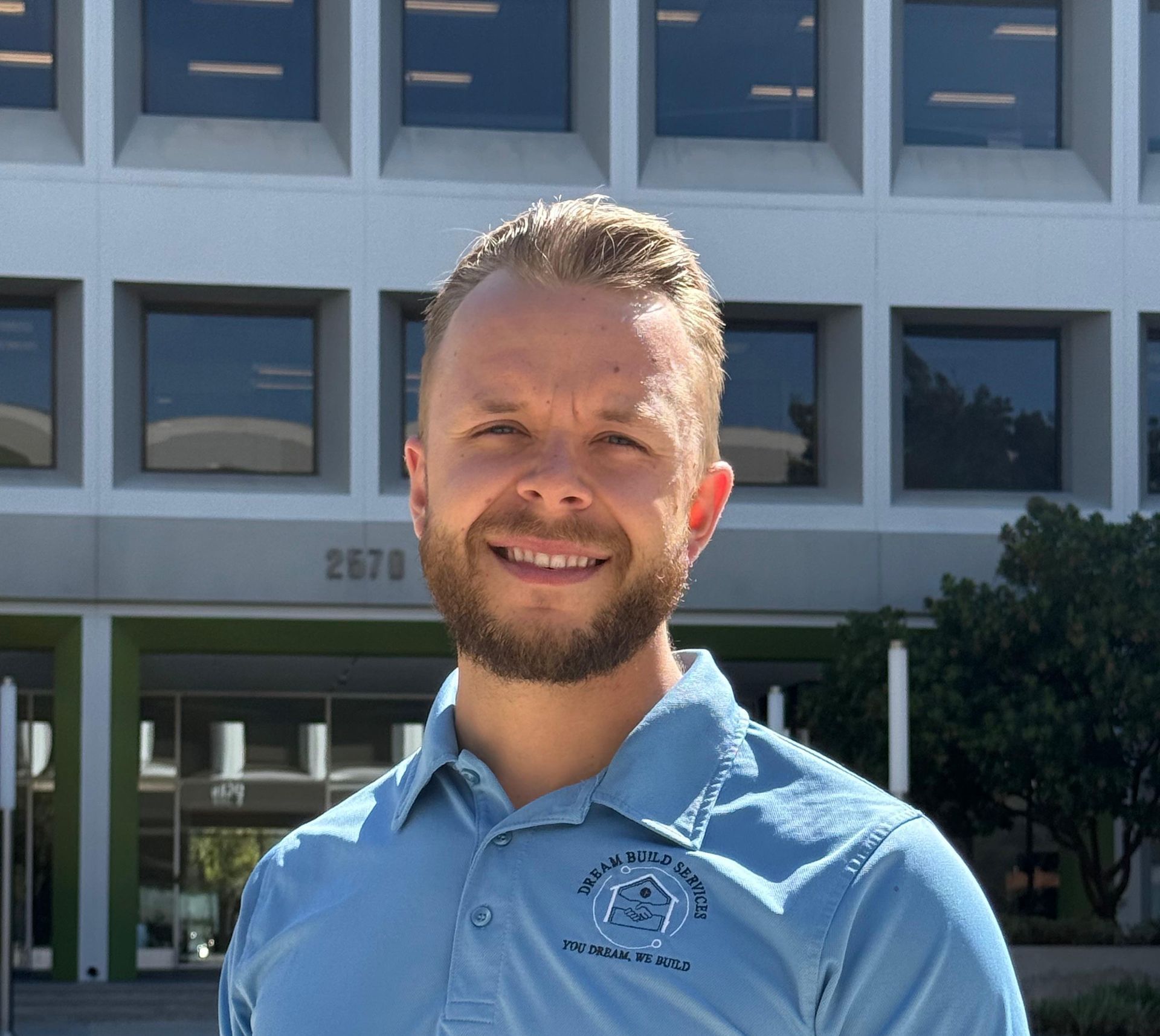 Man with a beard smiles in front of a building. He wears a blue collared shirt, the building has the number 2670 above its entrance.