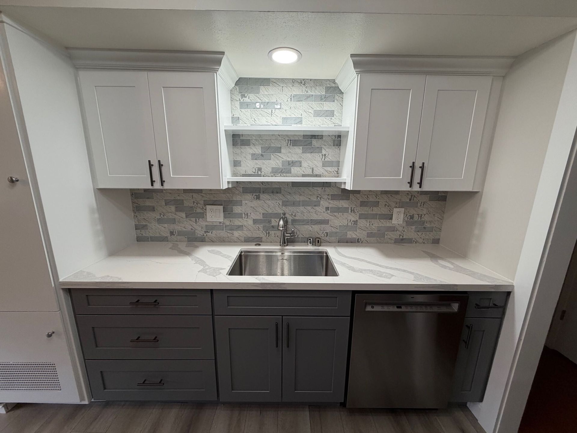 Modern kitchen remodel featuring gray shaker-style lower cabinets, white upper cabinets, quartz countertops with subtle veining, stainless steel appliances, built-in oven, sleek black cooktop, contemporary range hood, and textured stone tile backsplash. Renovation completed by Dream Build Services, showcasing high-quality craftsmanship, modern design, and functional layout for residential kitchen remodeling.