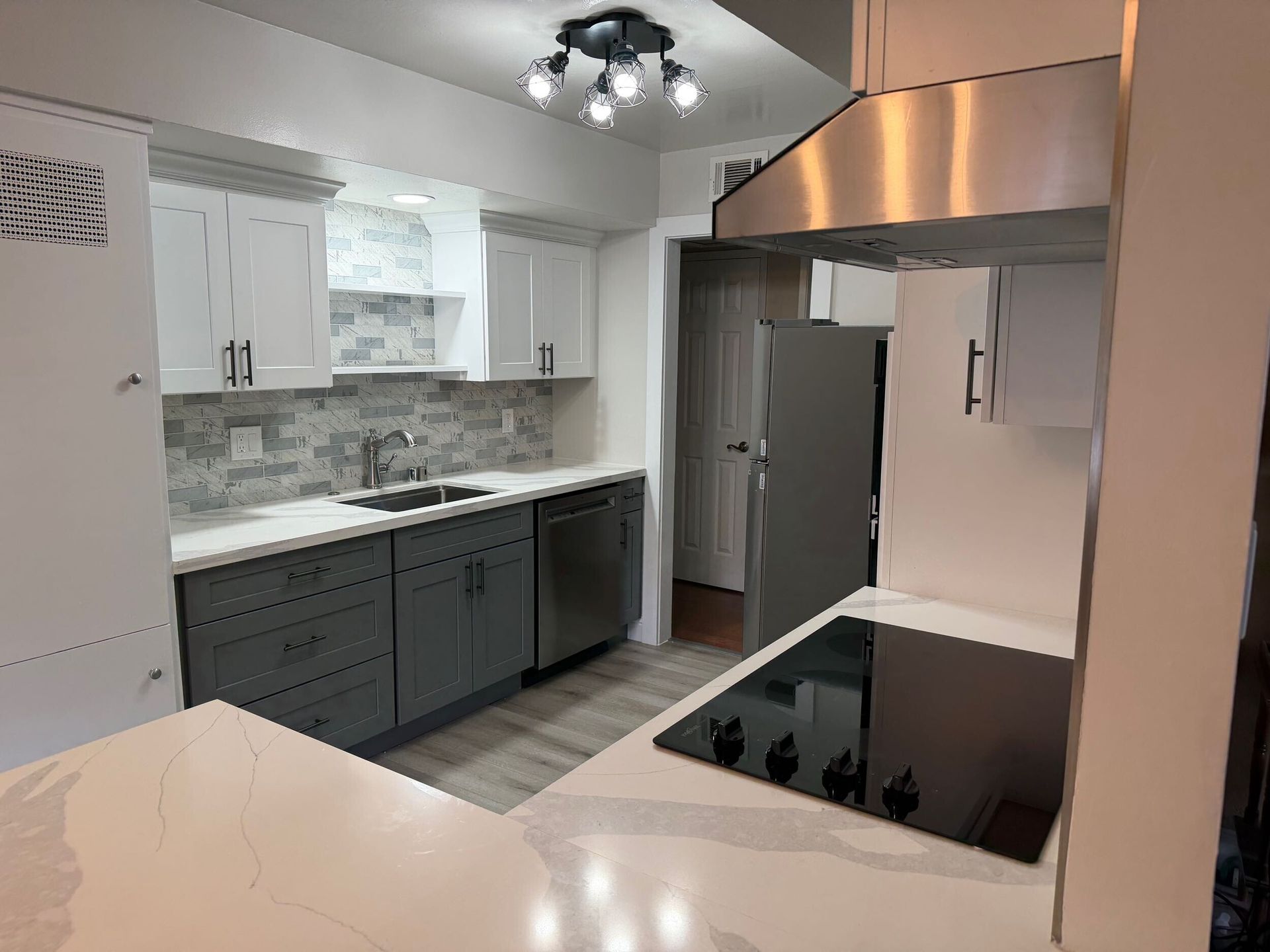 Modern kitchen remodel featuring gray shaker-style lower cabinets, white upper cabinets, quartz countertops with subtle veining, stainless steel appliances, built-in oven, sleek black cooktop, contemporary range hood, and textured stone tile backsplash. Renovation completed by Dream Build Services, showcasing high-quality craftsmanship, modern design, and functional layout for residential kitchen remodeling.