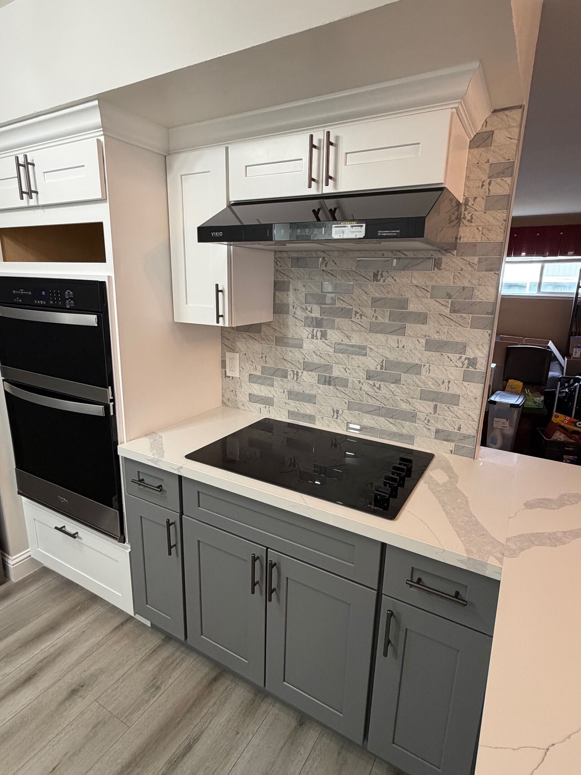 Modern kitchen remodel featuring gray shaker-style lower cabinets, white upper cabinets, quartz countertops with subtle veining, stainless steel appliances, built-in oven, sleek black cooktop, contemporary range hood, and textured stone tile backsplash. Renovation completed by Dream Build Services, showcasing high-quality craftsmanship, modern design, and functional layout for residential kitchen remodeling.