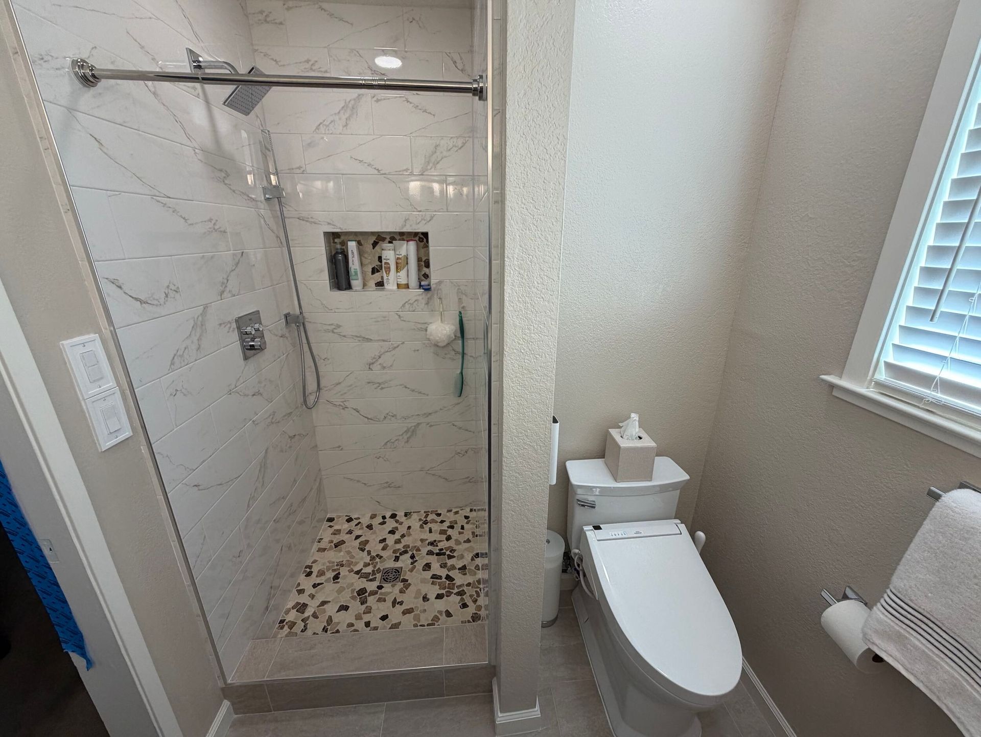 Updated bathroom remodel showcasing a modern walk-in shower with marble-style wall tile, pebble stone shower flooring, upgraded vanity with quartz countertop, contemporary sink and faucet, new lighting, and neutral color palette. Bathroom renovation completed by Dream Build Services, a residential remodeling contractor serving homeowners with bathroom and home renovation projects in the Bay Area.