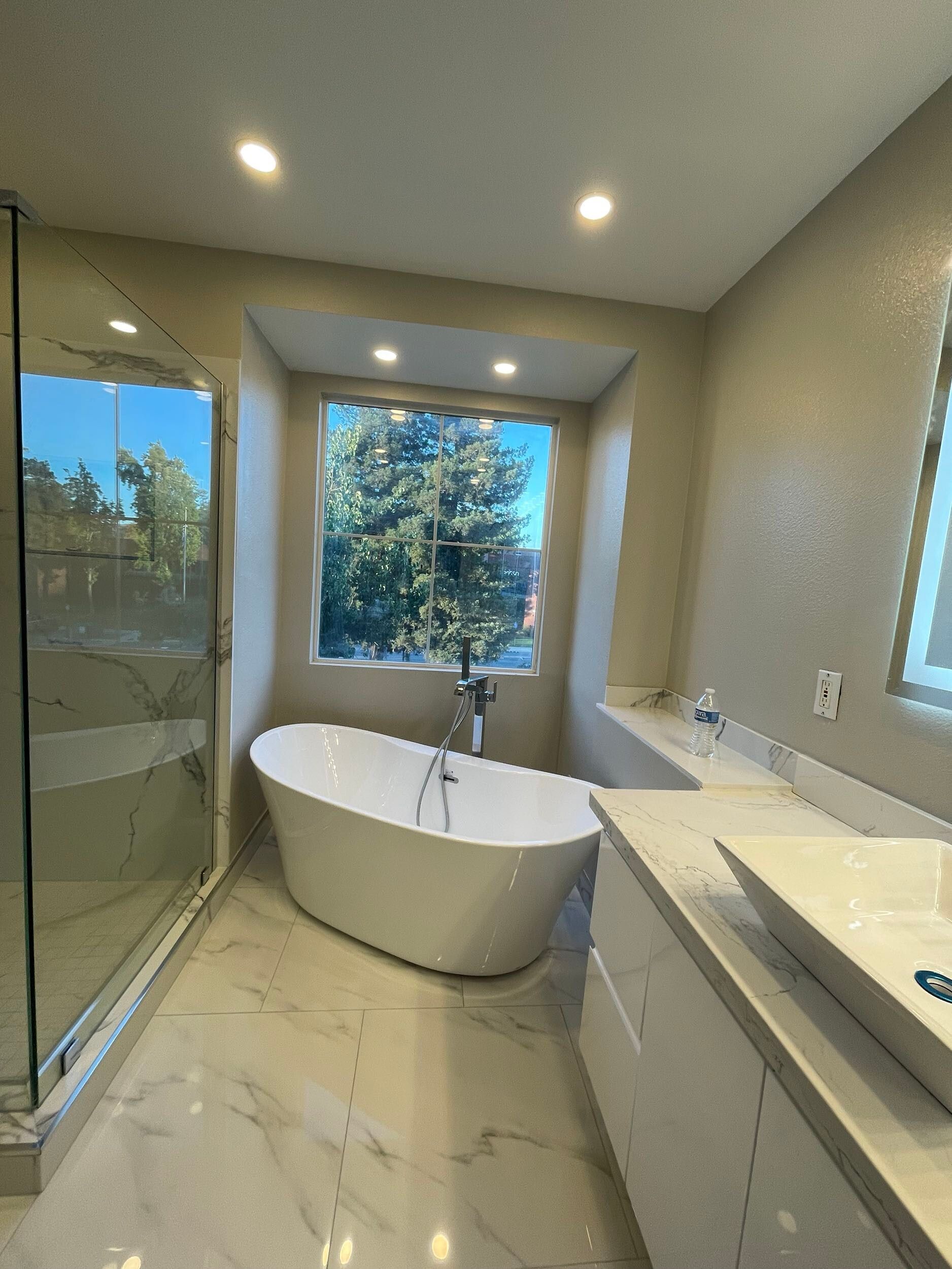 Luxury master bathroom remodel featuring a freestanding soaking tub by a large picture window, dual vessel sinks on a sleek white vanity, illuminated LED backlit mirror, recessed lighting, marble-look porcelain flooring, and frameless glass shower — custom design by Dream Build Services in the Bay Area.