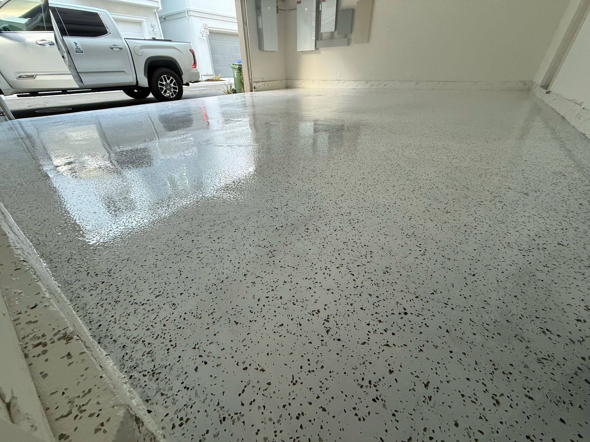 Glossy, speckled gray epoxy floor in a garage, reflecting the light and a white truck parked outside.