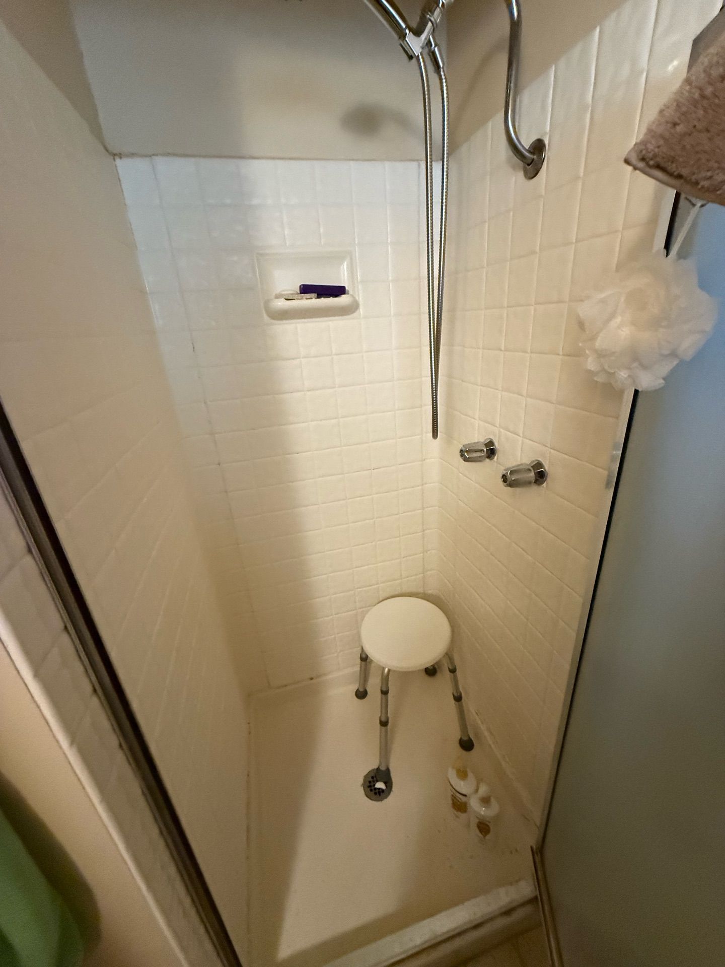 Small shower stall with white square tile walls and built-in soap niche, before bathroom remodel. Features a shower stool, handheld shower head, and simple white shower pan.