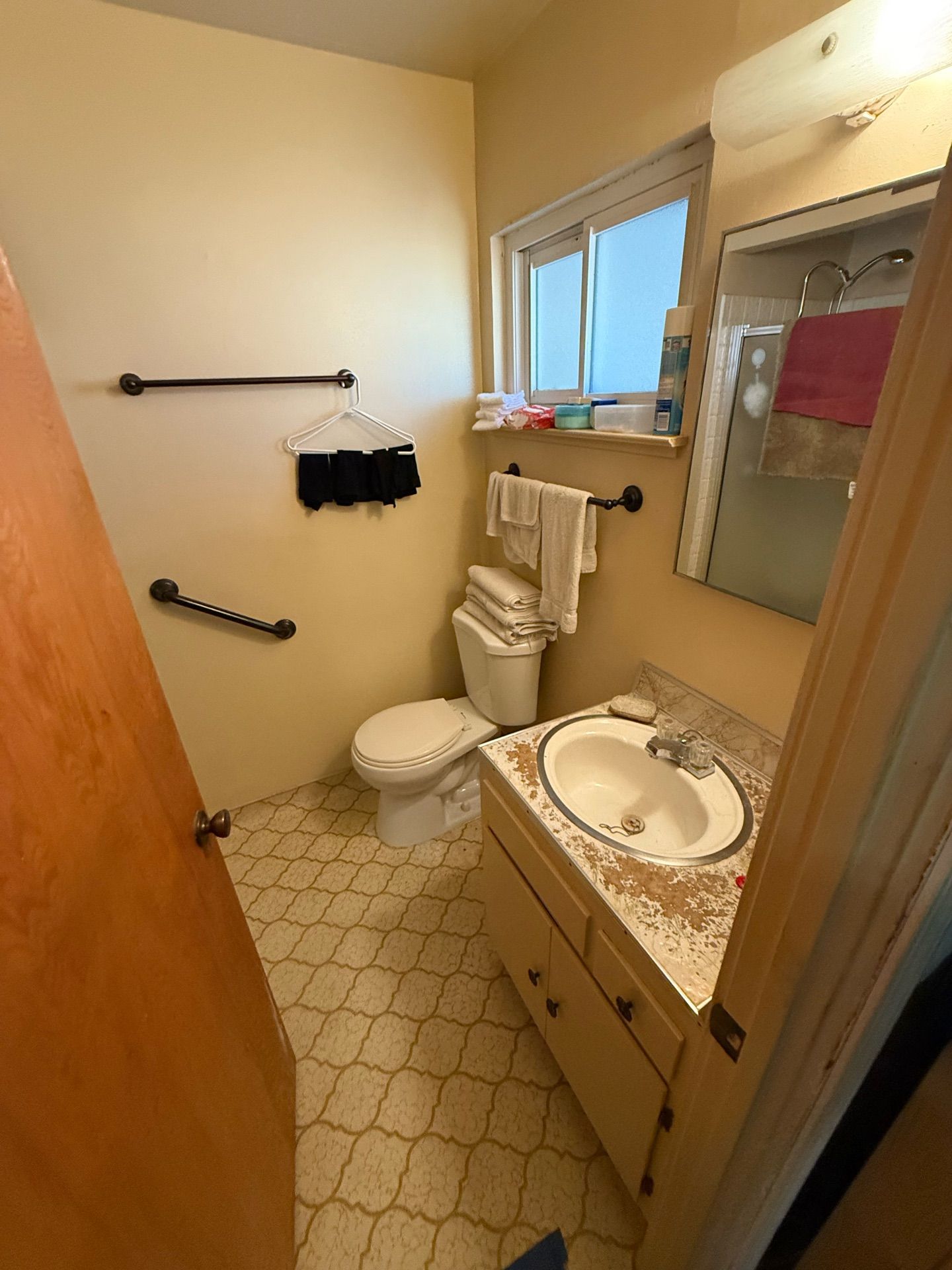 Outdated bathroom interior before remodel, with beige walls, patterned vinyl flooring, small vanity sink, wall-mounted mirror, and grab bars by the toilet.