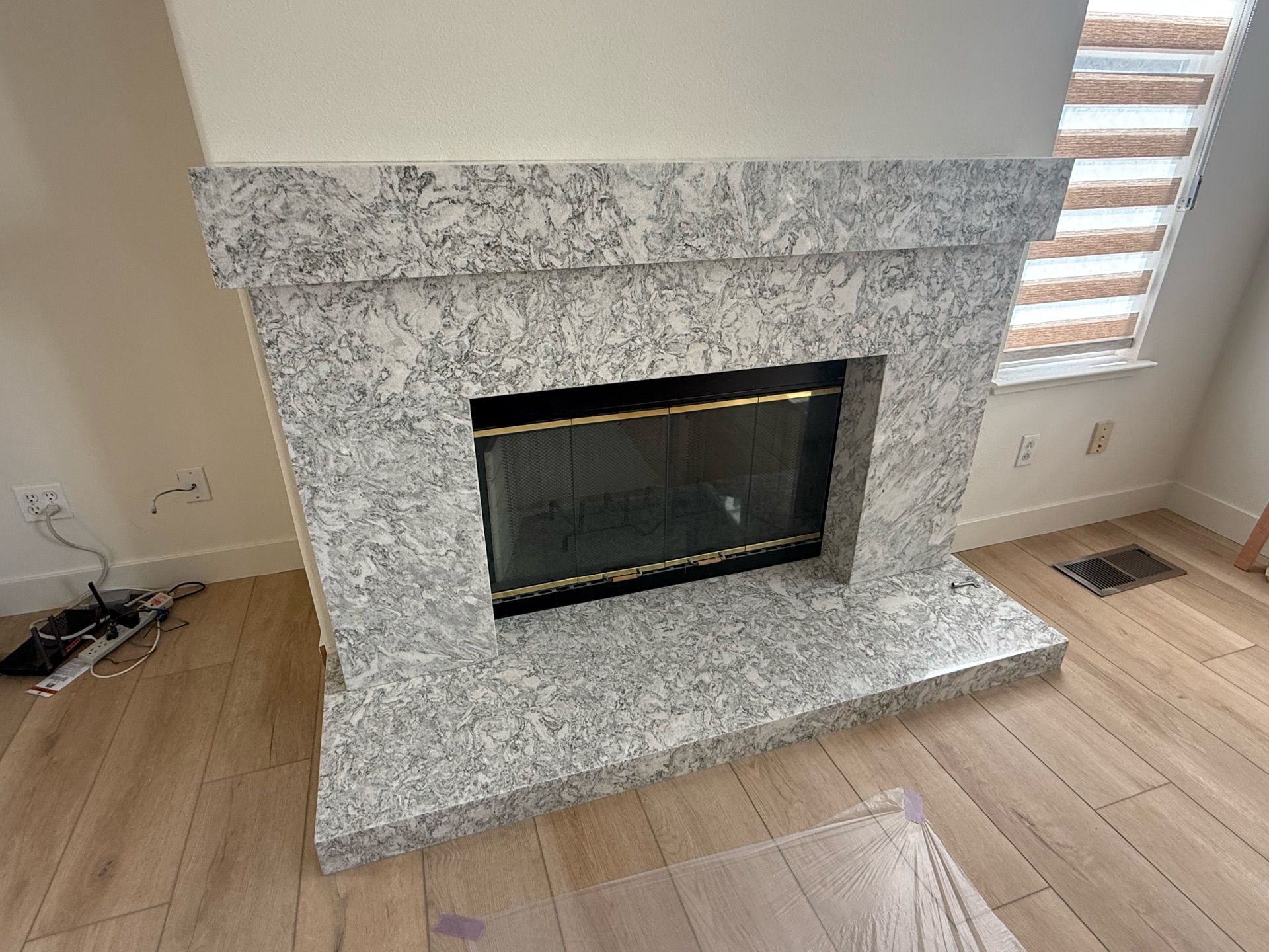 Granite fireplace with a black firebox and gold trim, set in a room with hardwood floors and a window with blinds.