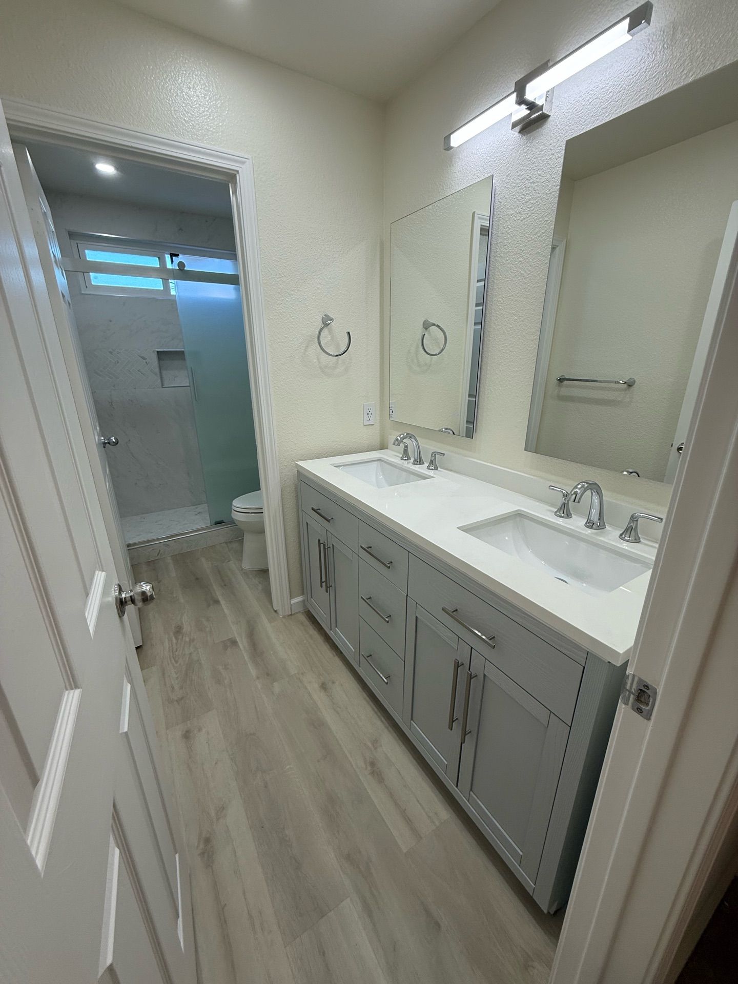 A light gray double-sink bathroom with a shower visible through the door. The vanity is also light gray, with a white countertop and silver fixtures.