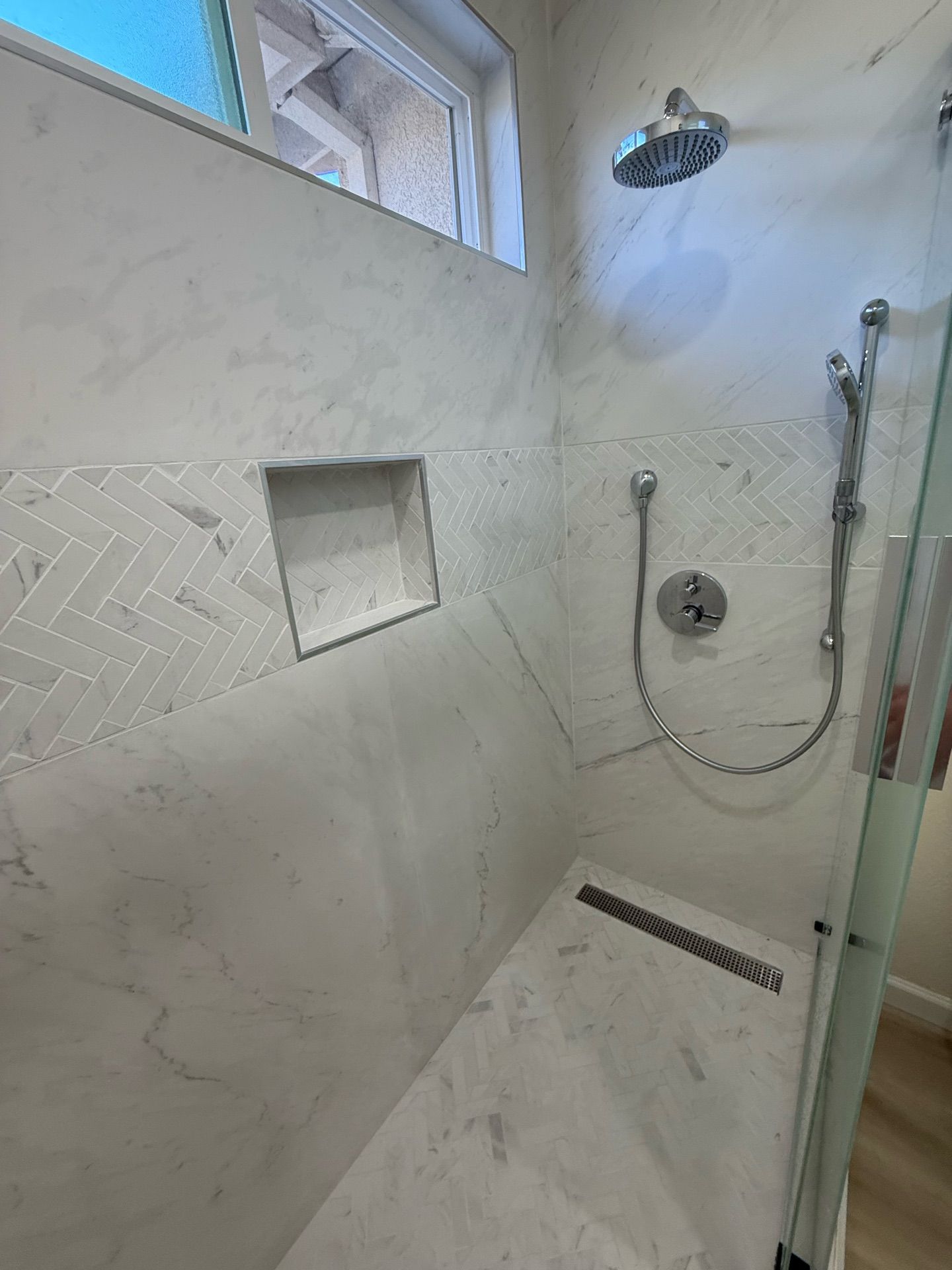 White marble shower with a built-in bench, rectangular niche, and herringbone accent tile; a glass door is visible.