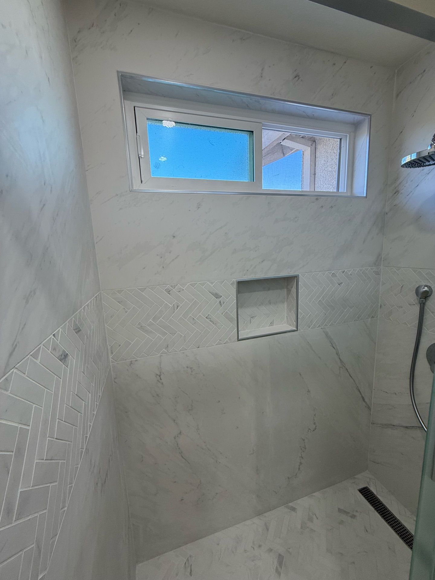 A newly tiled shower with a small window and built-in shelf. The walls and floor are white marble.