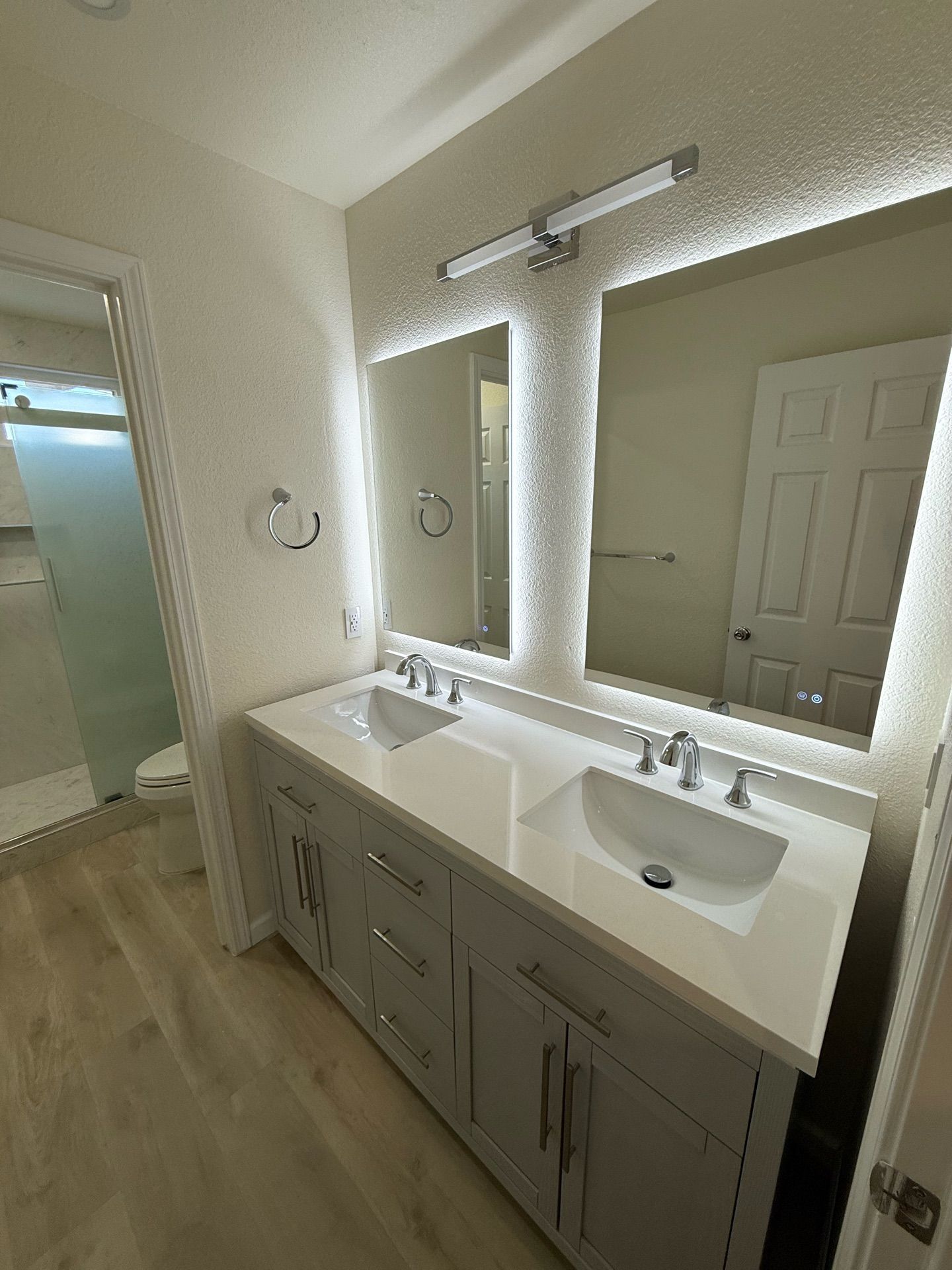 Bathroom with a double vanity, mirrors with LED lighting, and a glass-enclosed shower. The vanity is gray, and the flooring is light wood.