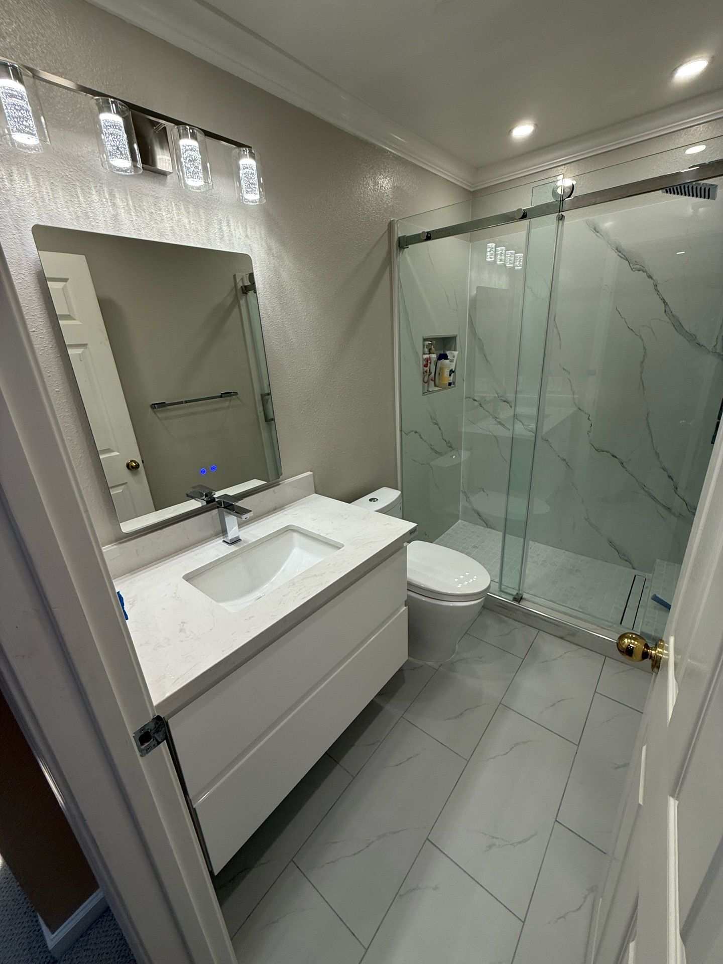 Contemporary bathroom remodel featuring a sleek white floating vanity with under-mount sink, frameless mirror with modern LED lighting, walk-in shower with glass sliding doors and marble-look porcelain tiles, and gray floor tiles with subtle veining — project by Dream Build Services in the Bay Area.