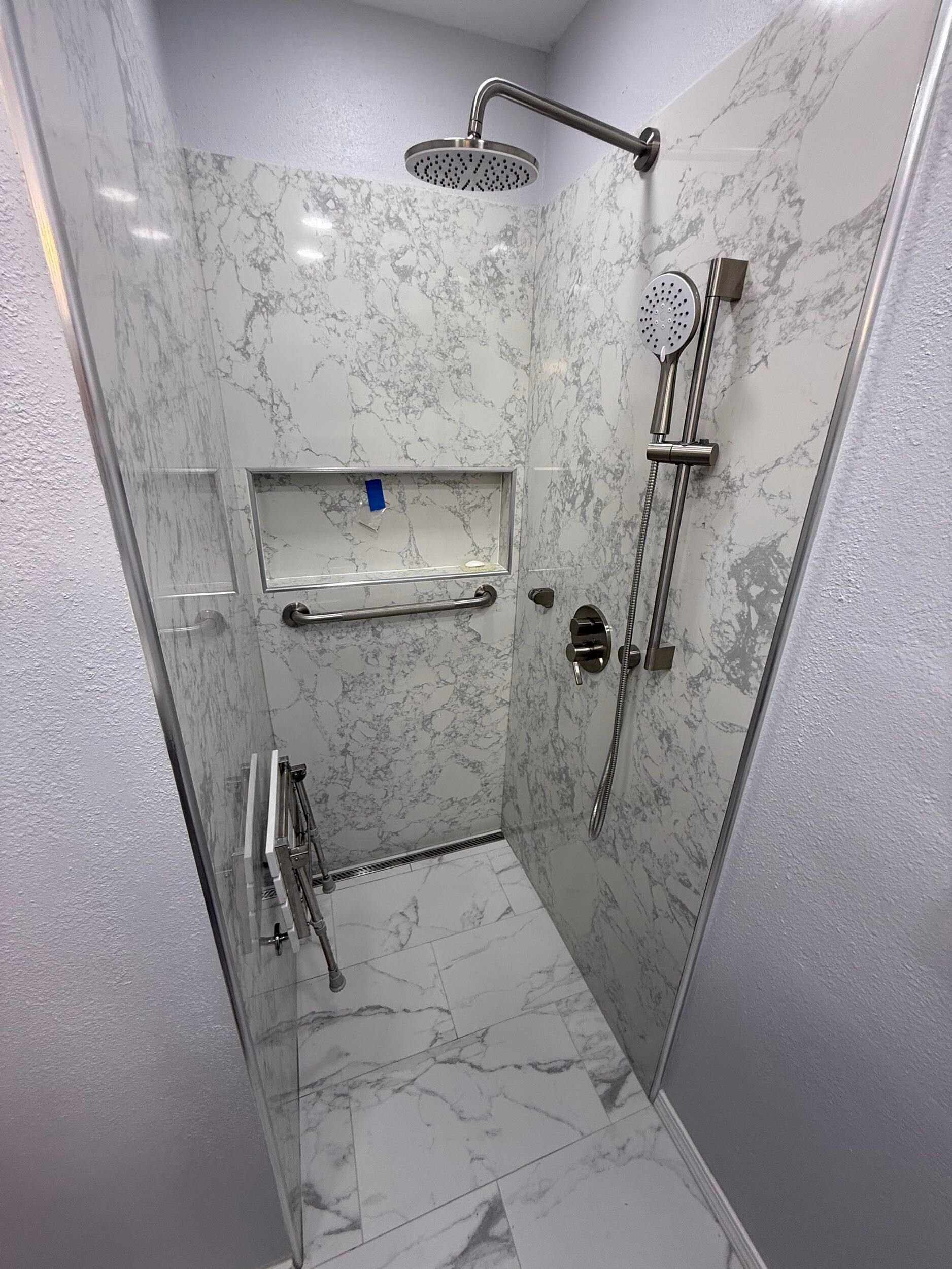 Modern walk-in shower after remodel, featuring white marble-look tiles, rainfall shower head, handheld shower, built-in niche, and sleek metal grab bars.