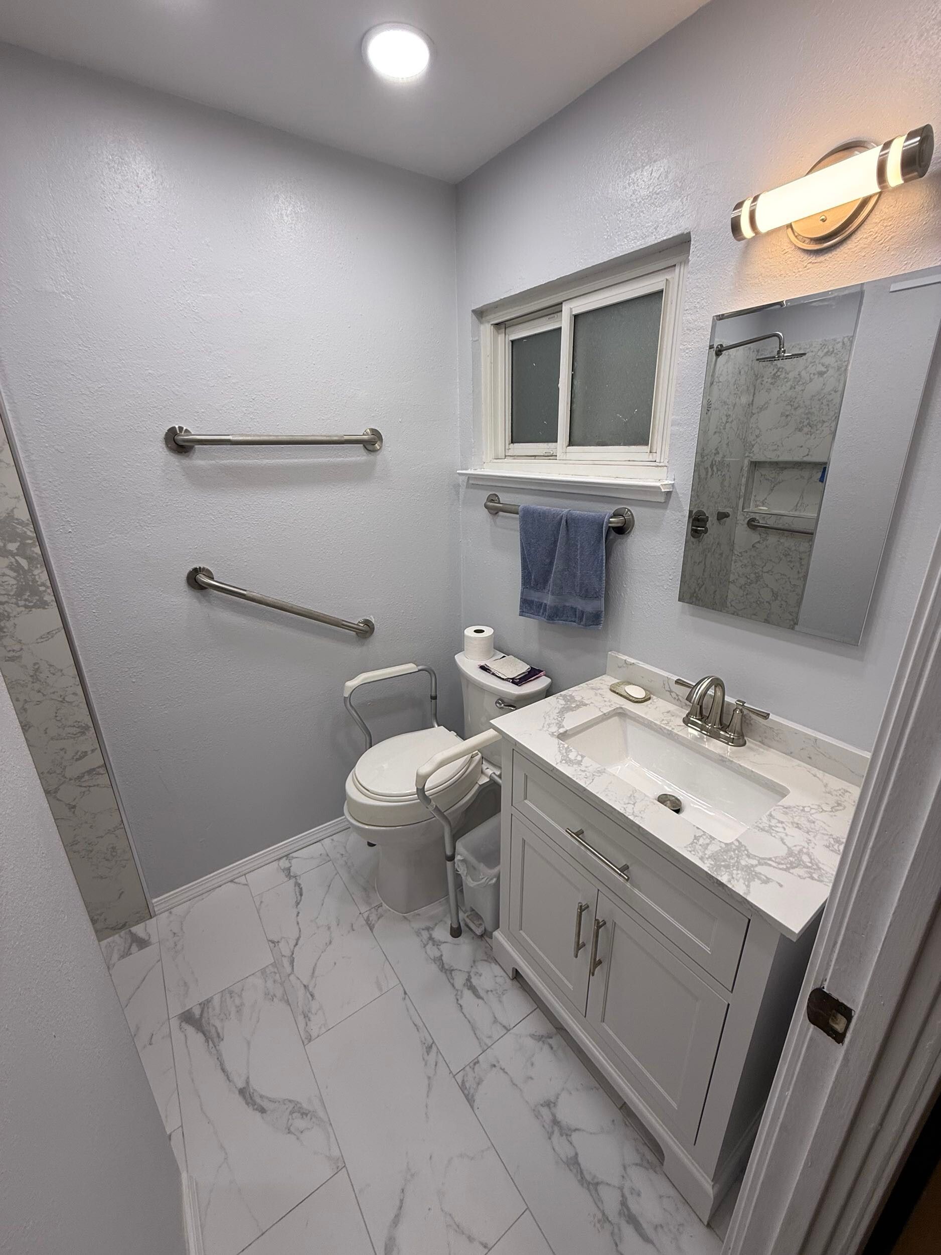 Newly remodeled bathroom with white vanity, marble countertop, updated lighting, gray walls, and matching marble-look floor and shower tiles for a clean, contemporary design.