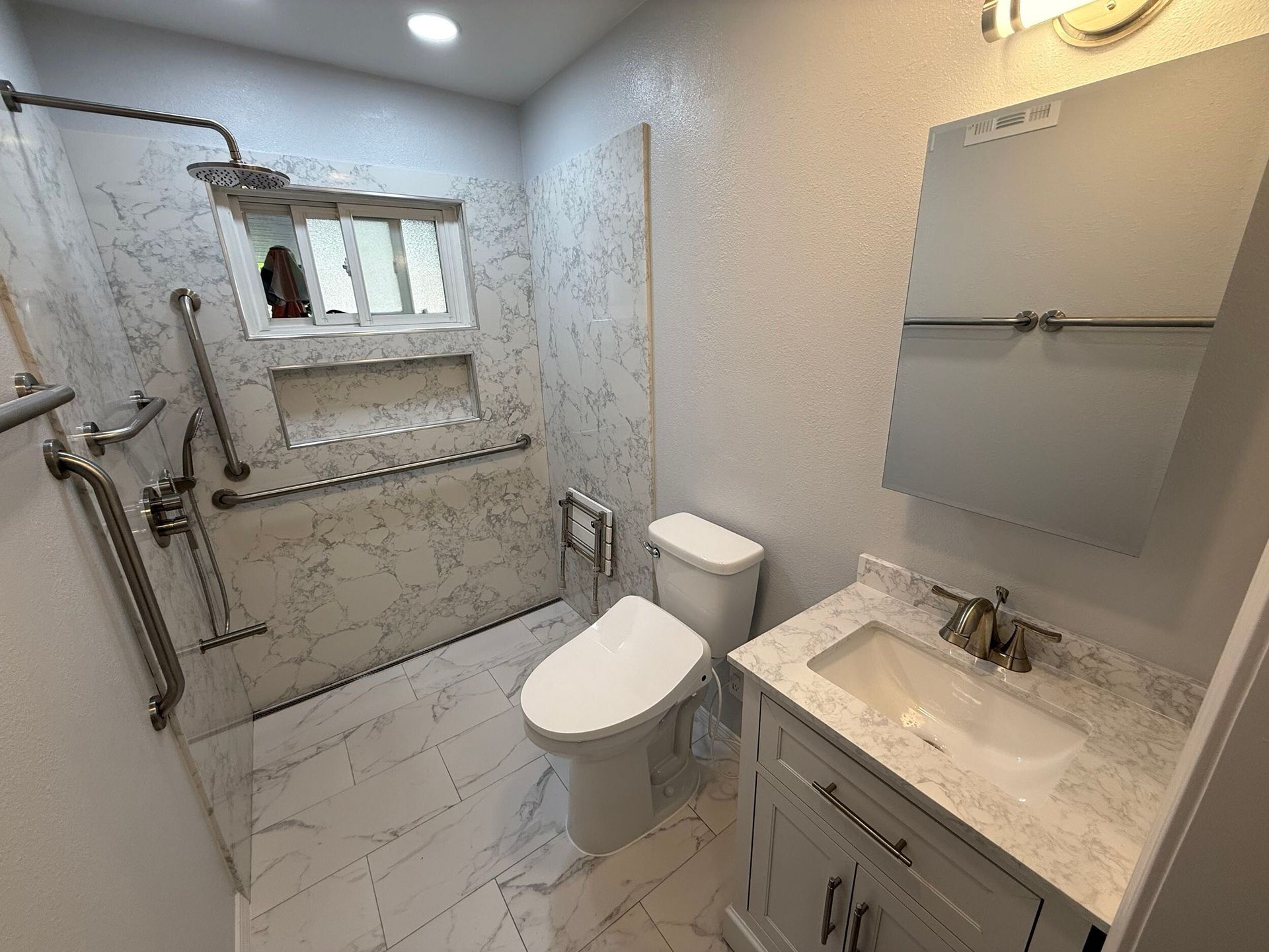 Modern bathroom remodel featuring white marble-style porcelain tiles on the floor and shower walls, walk-in shower with dual shower heads and accessibility grab bars, white vanity with under-mount sink and brushed nickel fixtures, completed by Dream Build Services in the Bay Area.