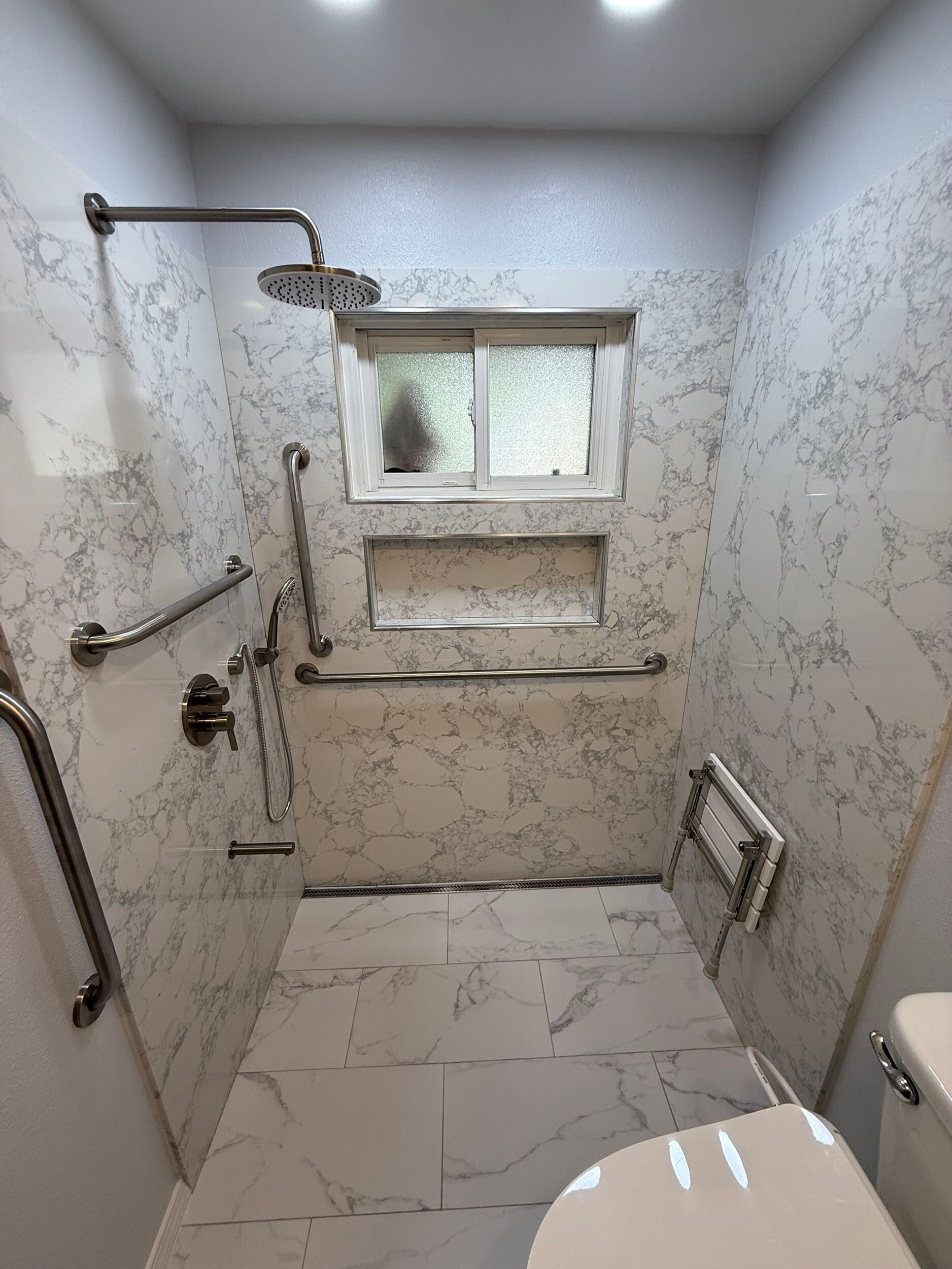 Modern bathroom remodel featuring white marble-style porcelain tiles on the floor and shower walls, walk-in shower with dual shower heads and accessibility grab bars, white vanity with under-mount sink and brushed nickel fixtures, completed by Dream Build Services in the Bay Area.