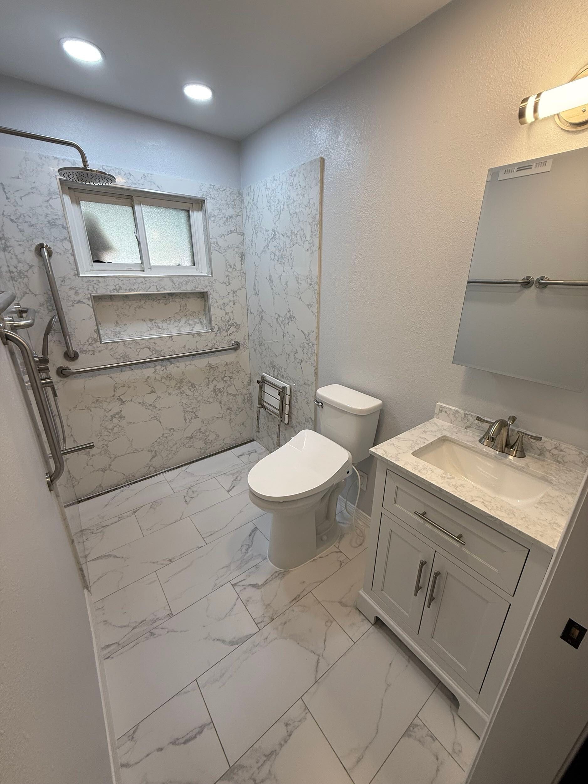 Modern bathroom remodel featuring white marble-style porcelain tiles on the floor and shower walls, walk-in shower with dual shower heads and accessibility grab bars, white vanity with under-mount sink and brushed nickel fixtures, completed by Dream Build Services in the Bay Area.