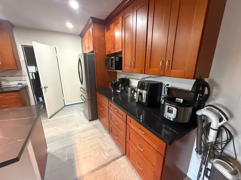 A kitchen with wooden cabinets , a refrigerator , a microwave , and a vacuum cleaner.