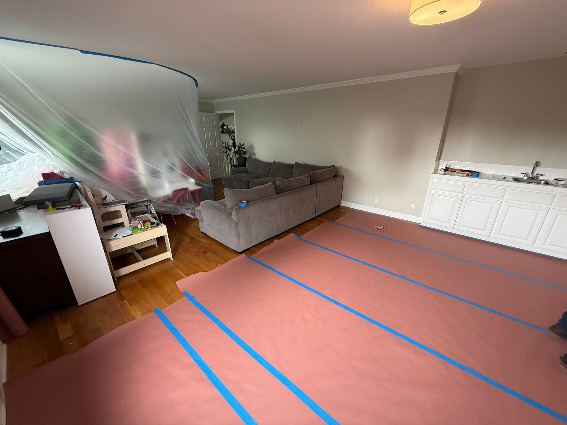 Living room undergoing renovations: pink flooring protection taped down, plastic sheeting covers a window and furniture.