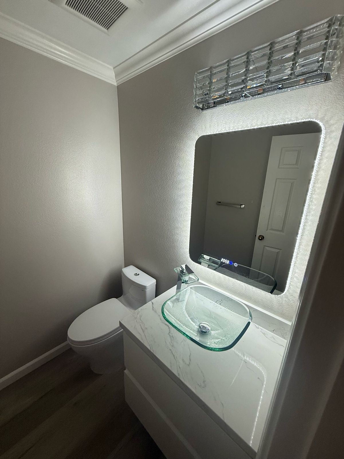 Modern bathroom remodel with wall-mounted LED mirror, glass vessel sink, and marble vanity top in a compact powder room.