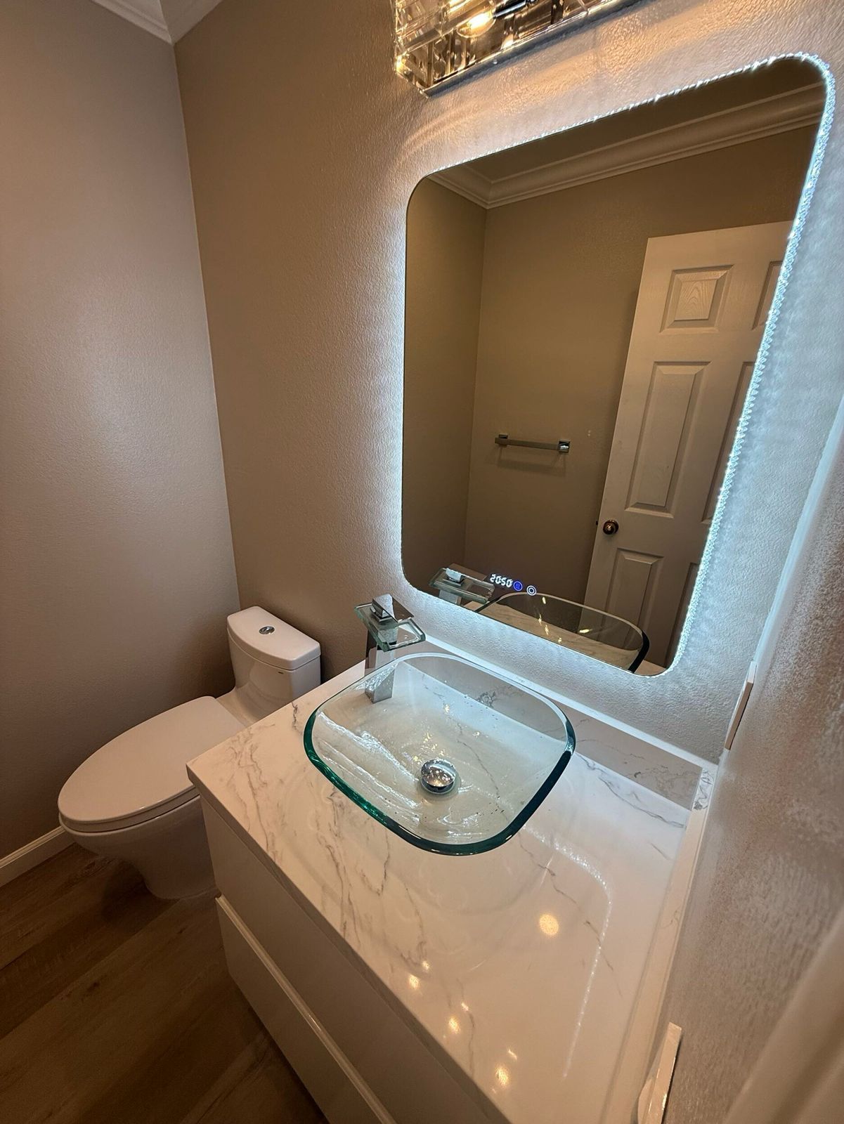 Modern bathroom remodel with wall-mounted LED mirror, glass vessel sink, and marble vanity top in a compact powder room.