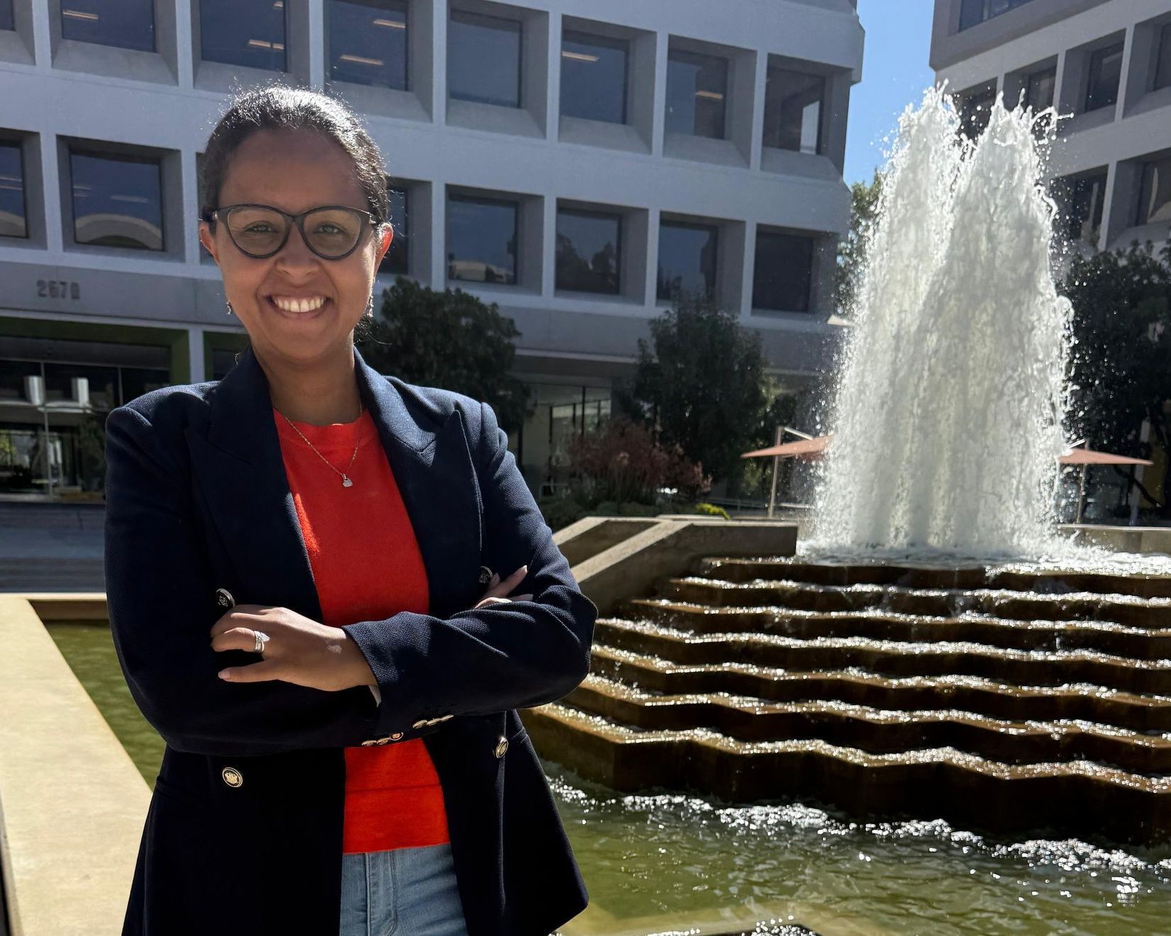Woman with crossed arms smiles in front of a fountain and building. She wears glasses, a red top, and a navy blazer.