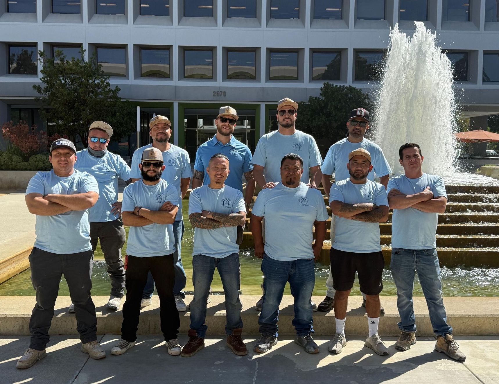 A group of men in light blue shirts stand in front of a fountain and building, arms crossed, looking at the camera.