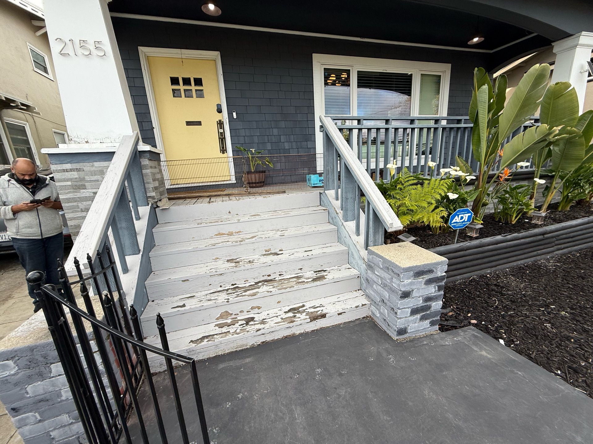 A man is standing in front of a blue house with a yellow door.
