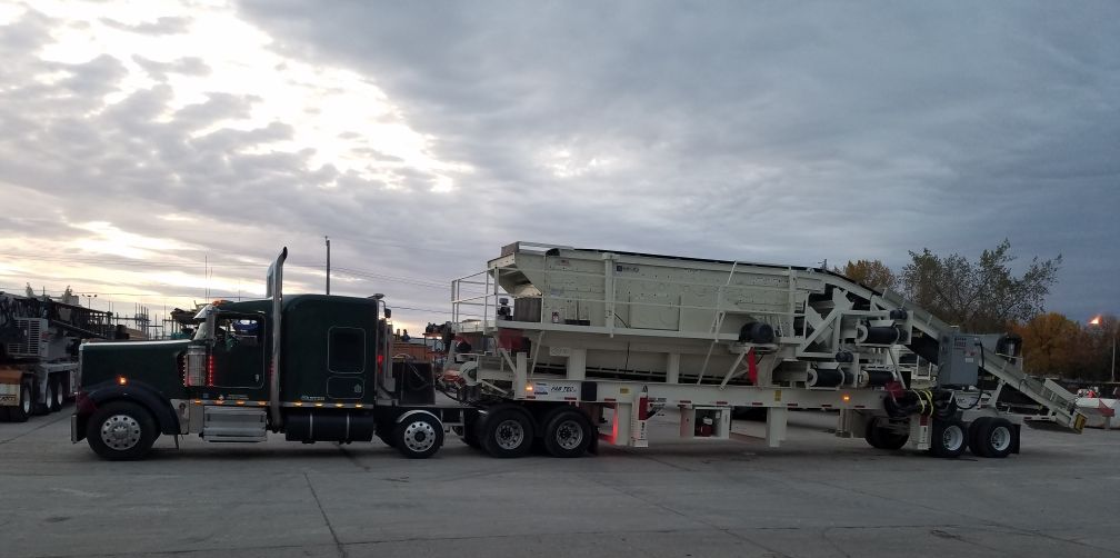 A black semi-truck hauls a large, white industrial machine on a flatbed trailer under a cloudy sky.