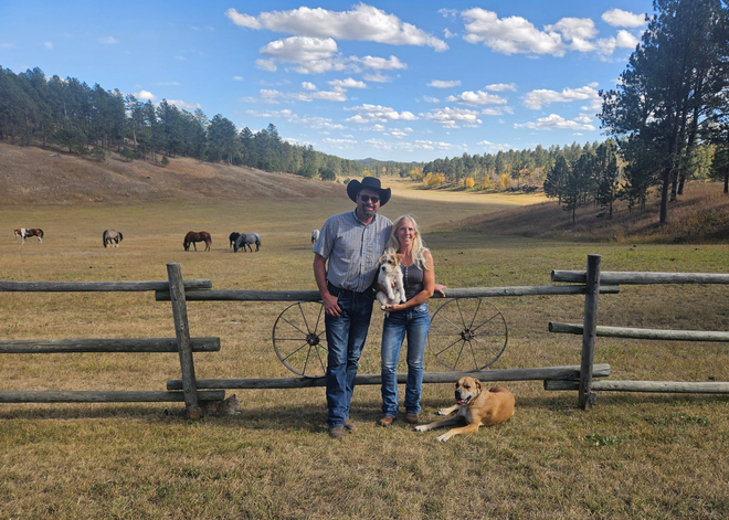 Rick and Sheila Carter from Rick Carter Trucking on horseback in a green field with dogs. Clear blue sky.