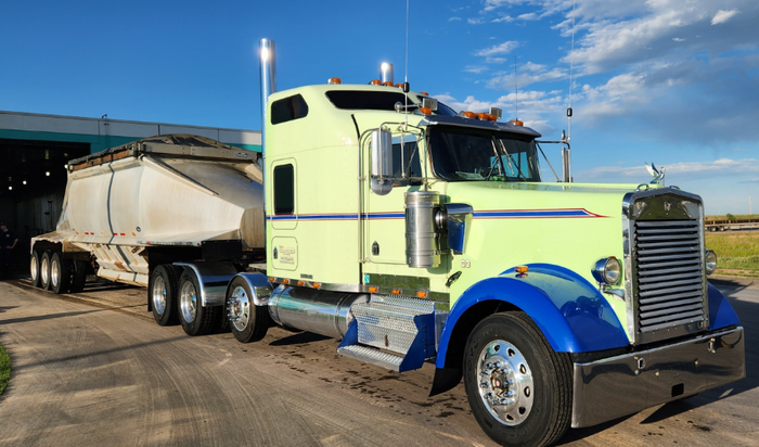 Light green semi-truck with a white trailer, blue fenders, parked under a sky with clouds.