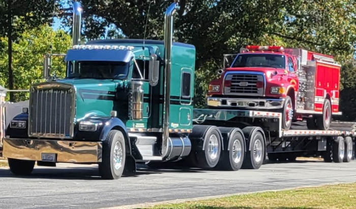 Green semi-truck towing a red fire truck on a flatbed trailer. Sunny day.