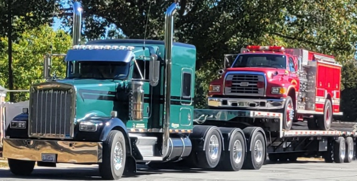 A semi-truck with a red and silver cab and a black trailer parked on a gravel lot under a blue sky.