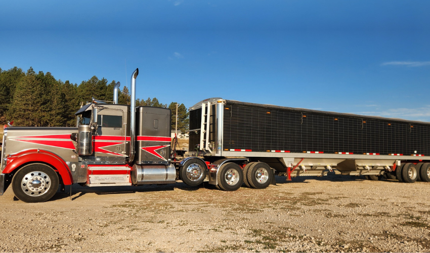 A semi-truck with a silver and red cab and a black cargo trailer parked on gravel under a blue sky.