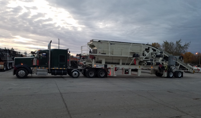 A semi-truck carrying a large white industrial machine on a flatbed trailer, parked on concrete.