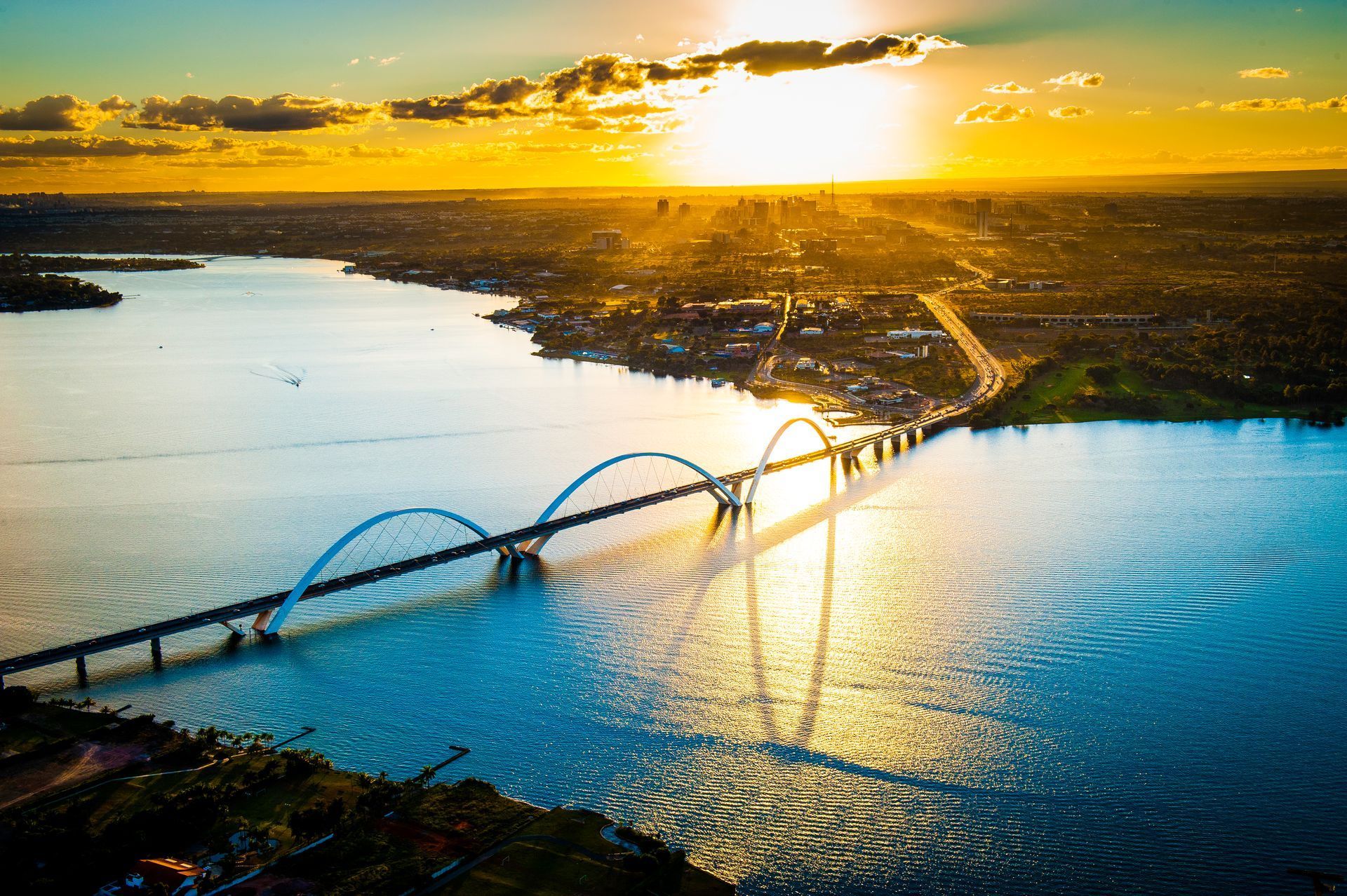 Pôr do sol sobre uma ponte com três arcos cruzando um rio; paisagem urbana ao fundo.