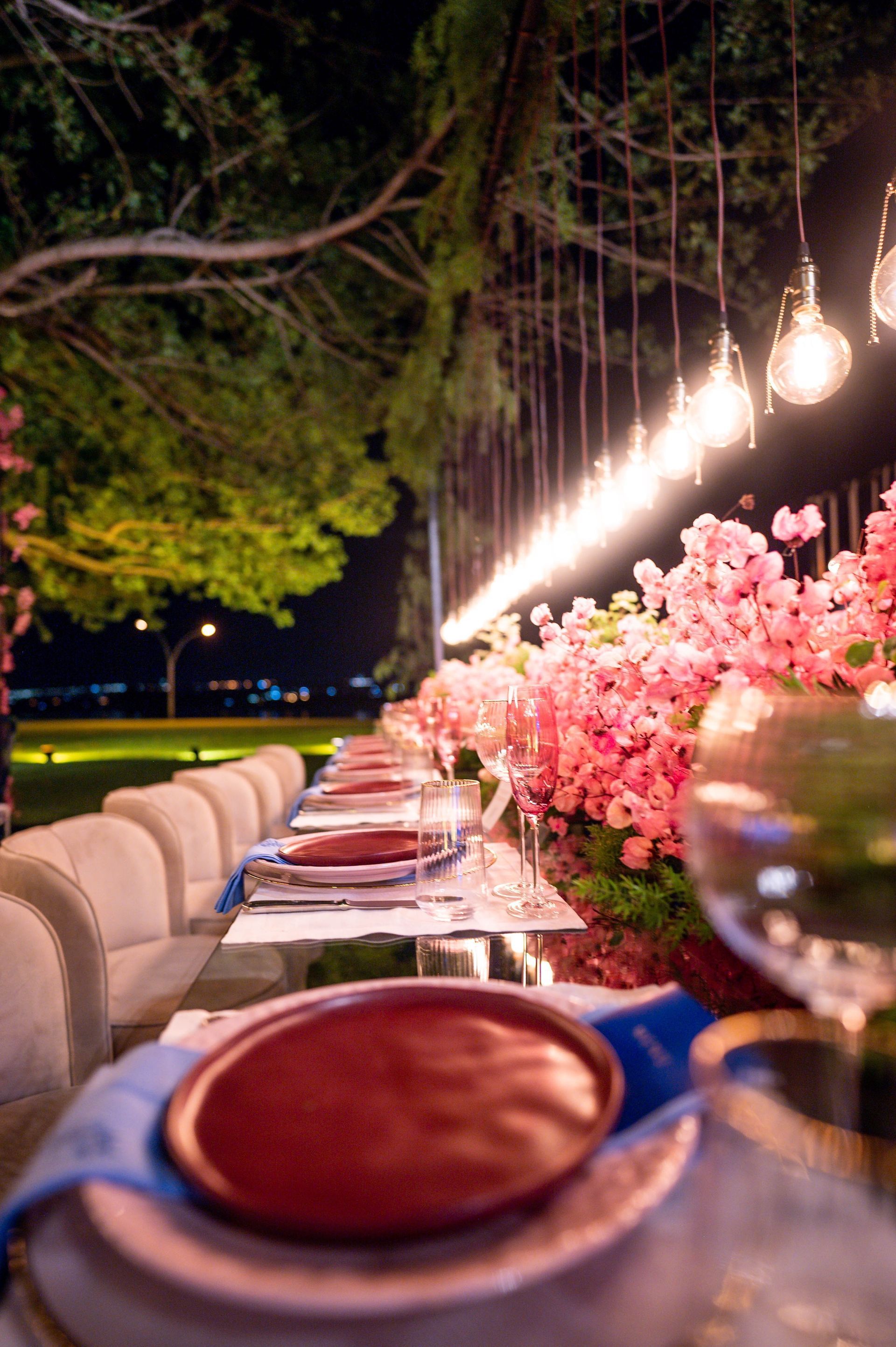 Mesa longa posta para um jantar ao ar livre à noite, iluminada por luzes penduradas e flores rosas.